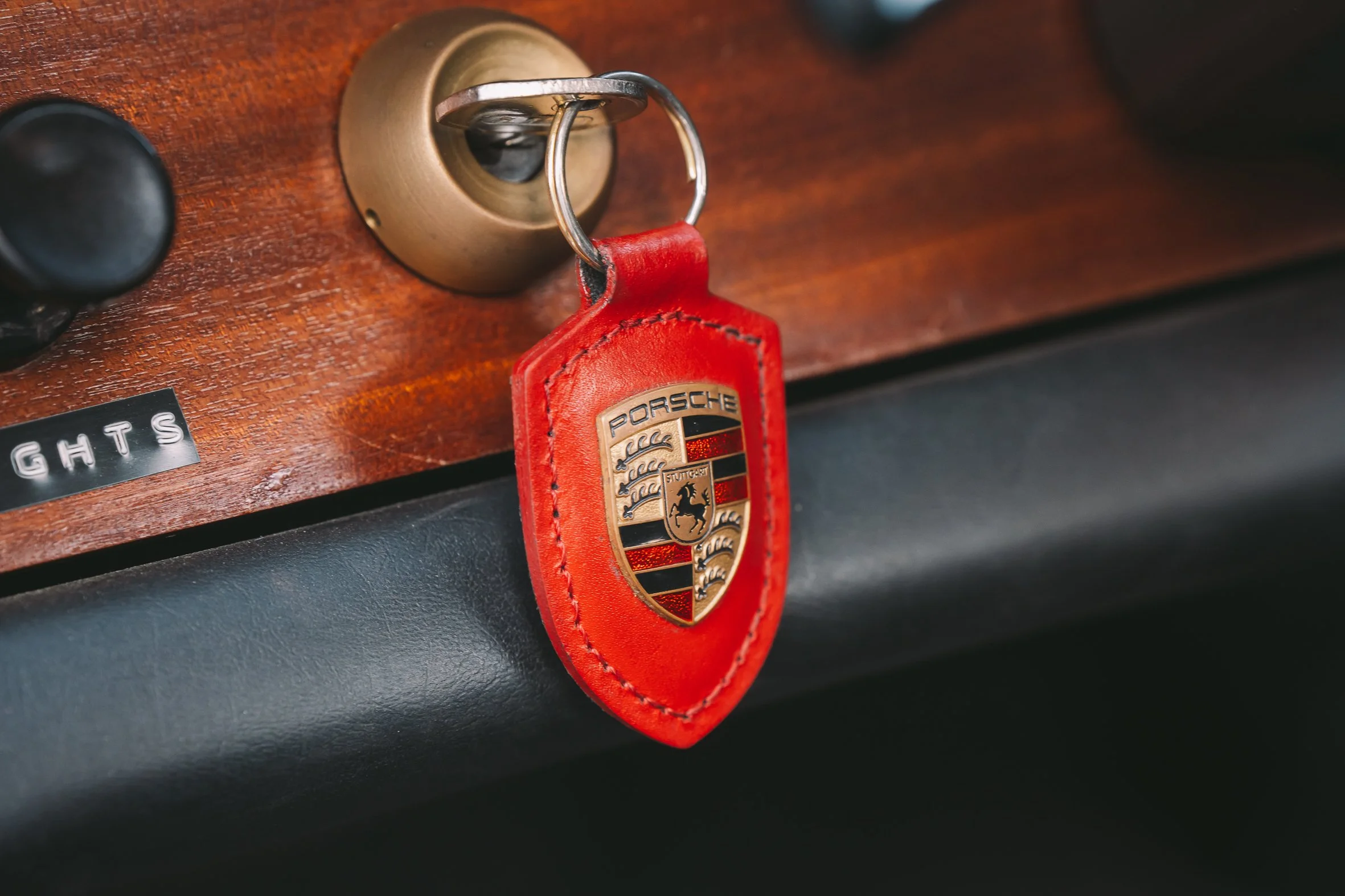 Close-up of a Porsche keychain hanging from a brass keyhole on a wooden panel in a car interior.