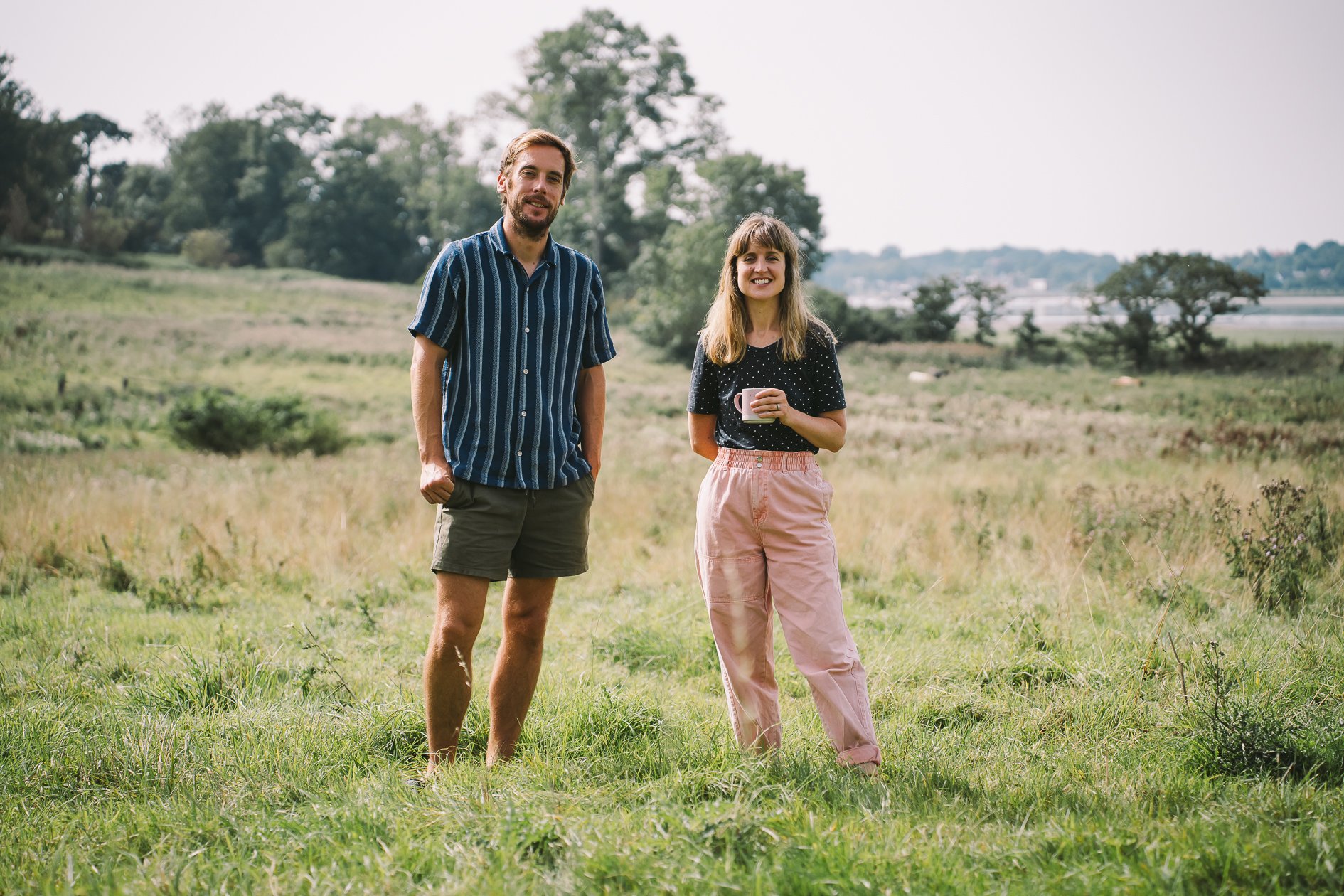 A man and woman standing outdoors in a grassy field with trees in the background. The man is wearing a striped button-up shirt and shorts, and the woman is wearing a black polka-dot shirt and pink pants, holding a mug, smiling at the camera.