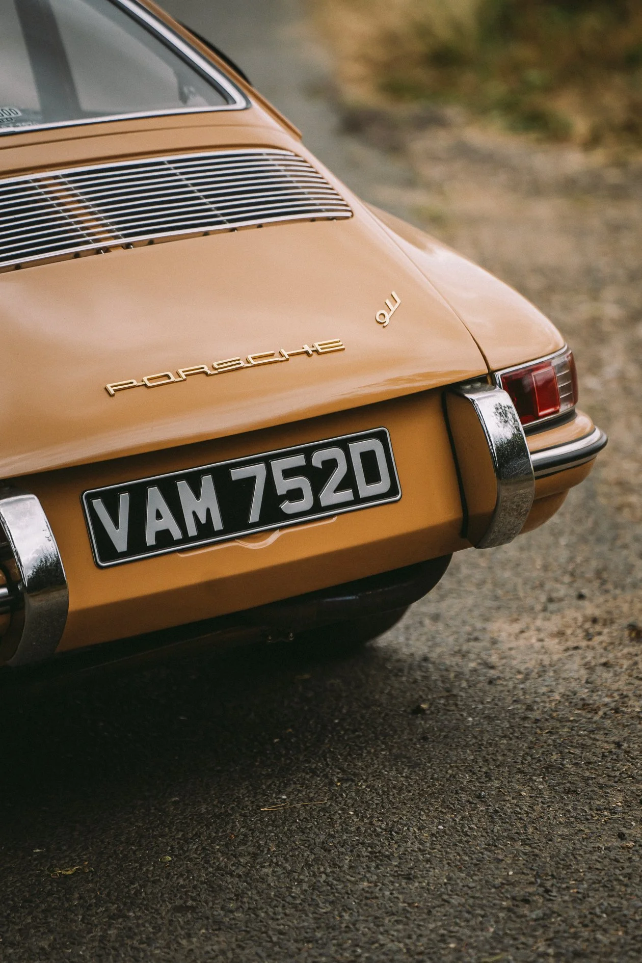 Close-up of the rear of a vintage gold Porsche 911 with a black and white license plate reading 'VAM 752D' and a chrome bumper.
