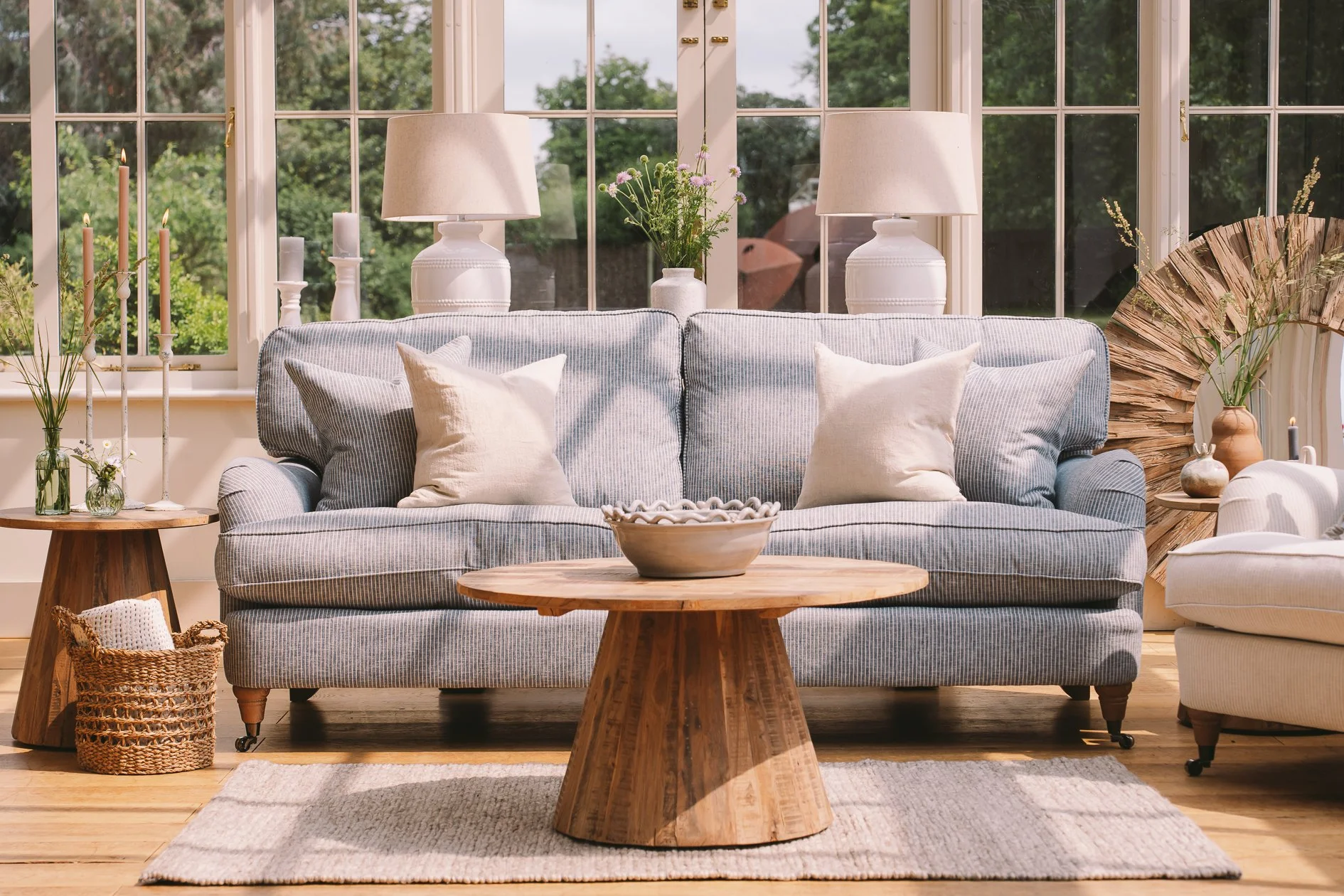 A cozy living room with a striped sofa adorned with white pillows, flanked by wooden side tables holding vases with flowers, in front of a large window with a scenic view of trees outside.