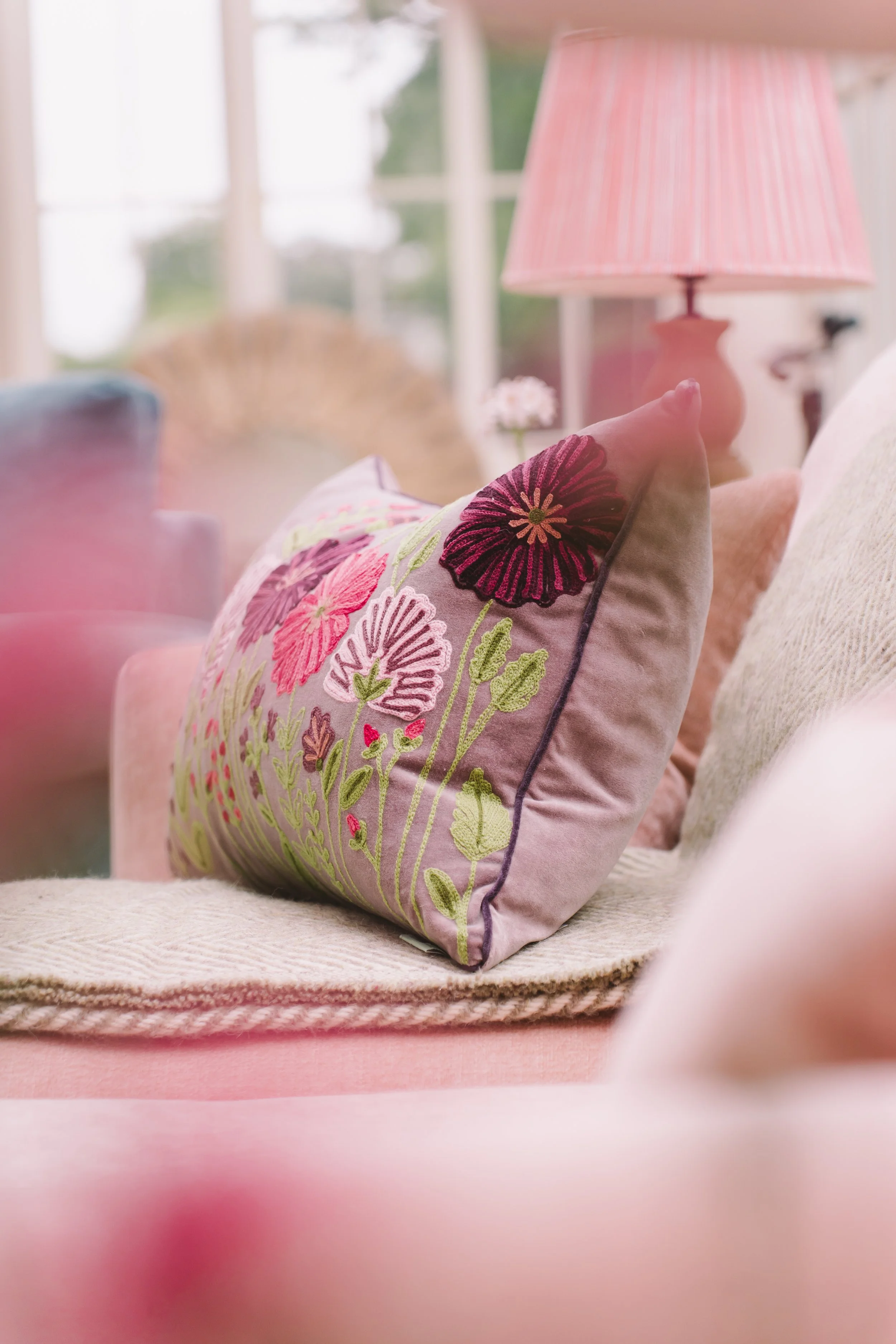 Close-up of a decorative embroidered pillow with pink, purple, and green floral design, resting on a beige sofa in a bright room with a pinkish lampshade and window in the background.