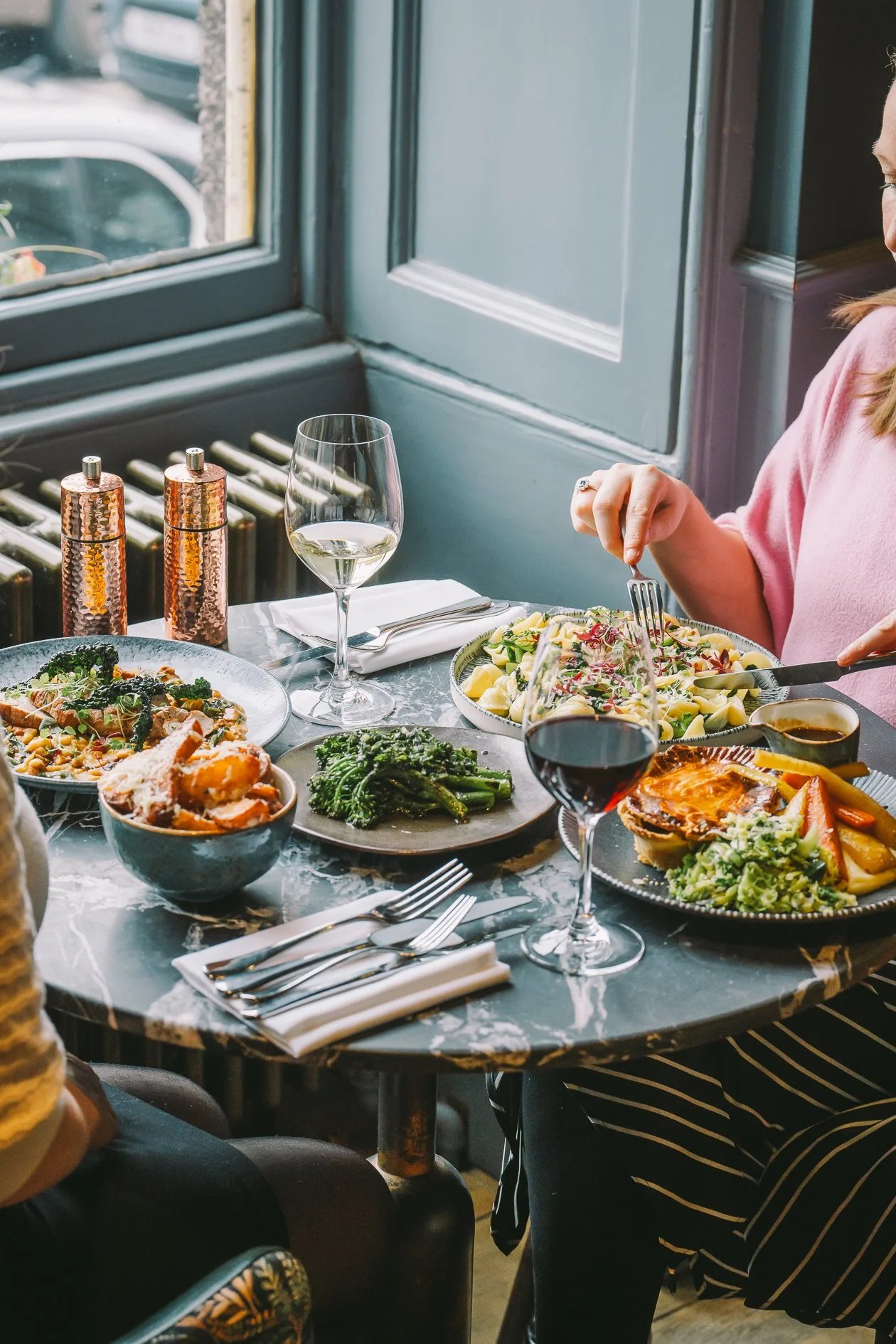 A table set with various dishes including salads, pasta, roasted vegetables, and a pie, with wine glasses and cutlery, near a window in a restaurant.