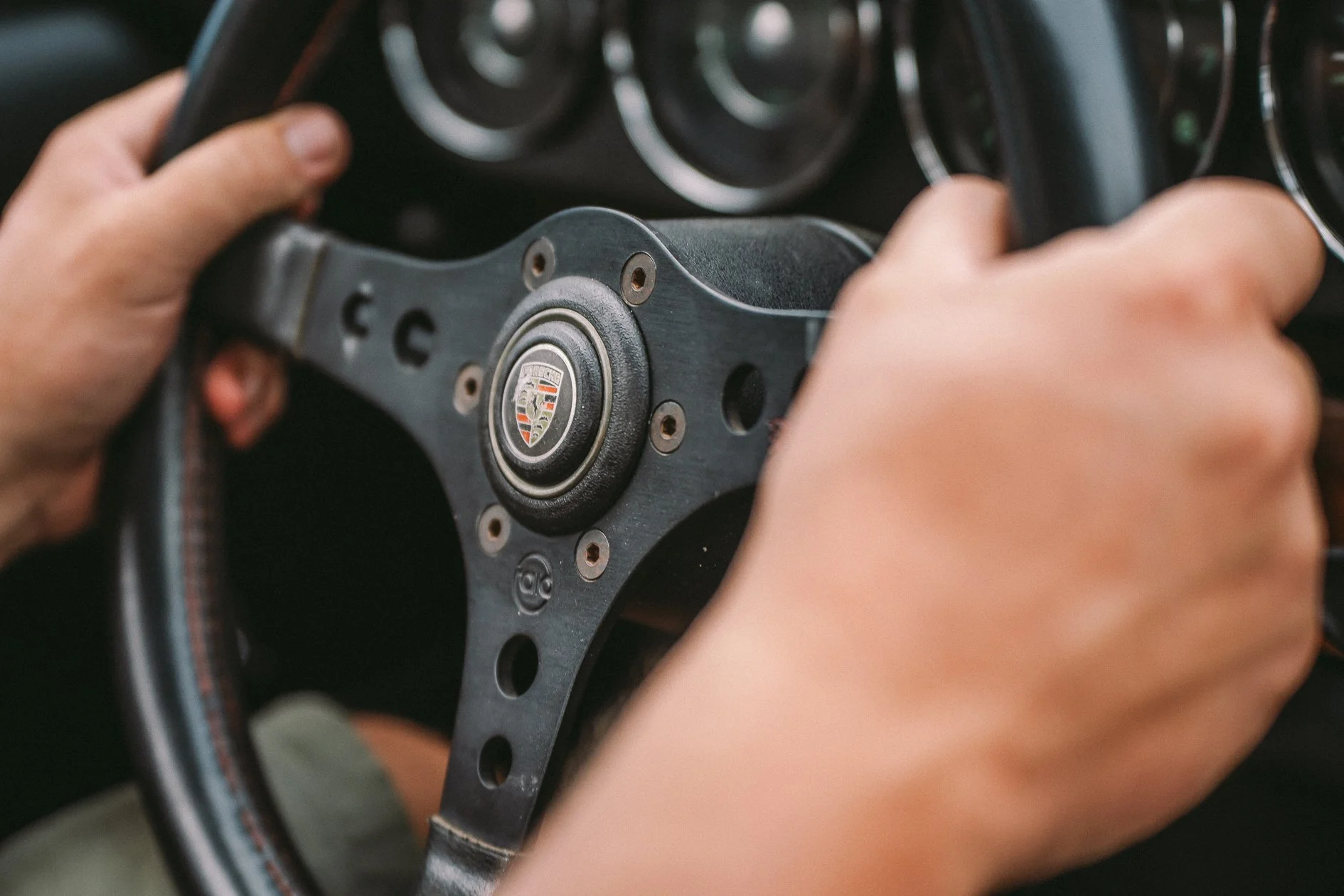 Close-up of a person holding a vintage Porsche steering wheel with both hands, focusing on the central hub with the Porsche logo.