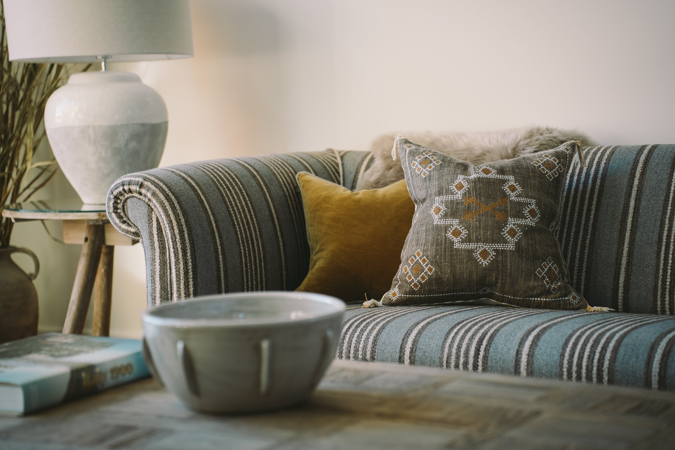 Living room corner with a striped sofa, decorative pillows, a large white ceramic lamp, and a side table with a plant in the background.