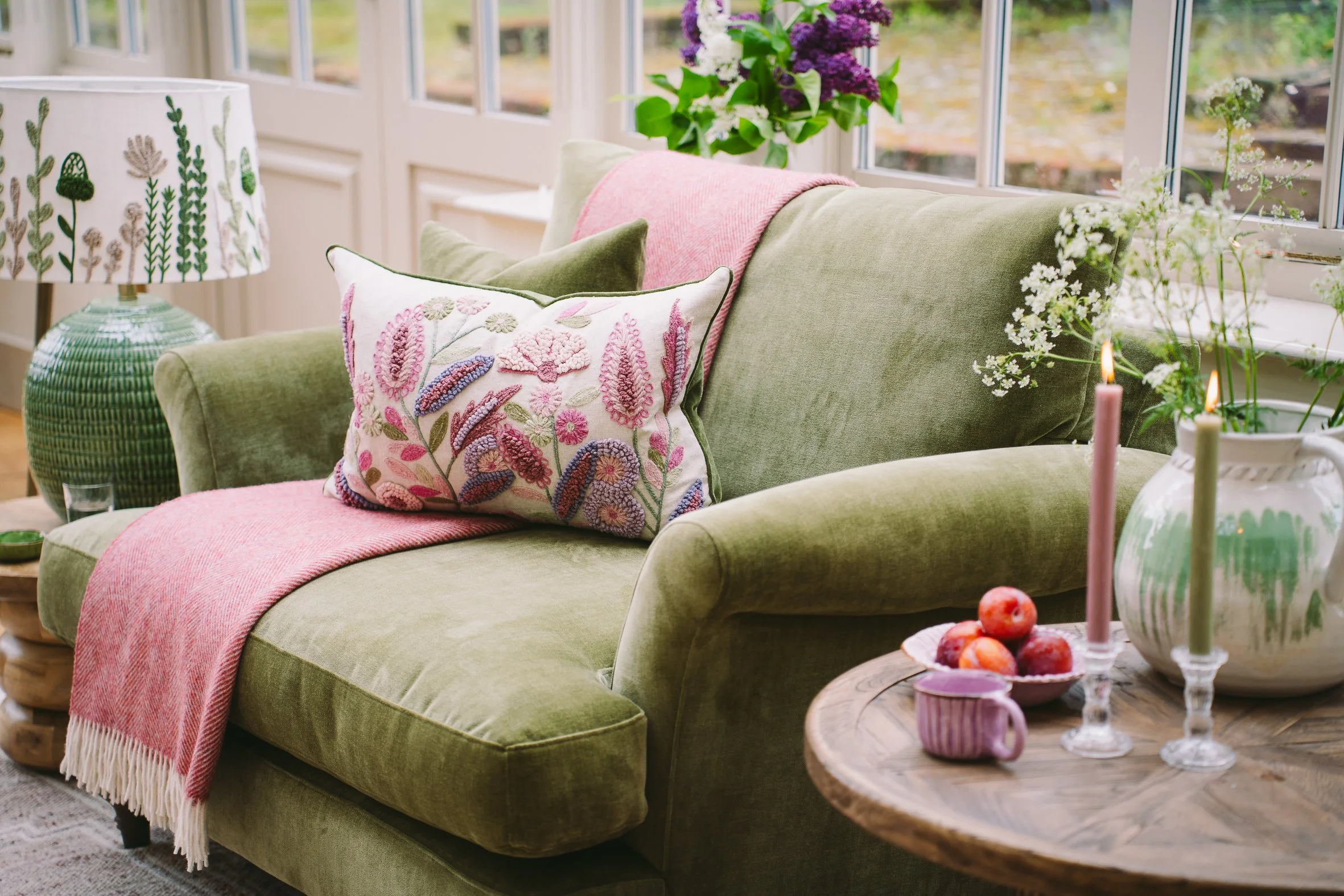 A green upholstered sofa with embroidered floral pillows, a pink throw blanket, floral decor, a lamp with a botanical pattern, a round wooden coffee table with candles and apples, and large windows with a view of a garden.