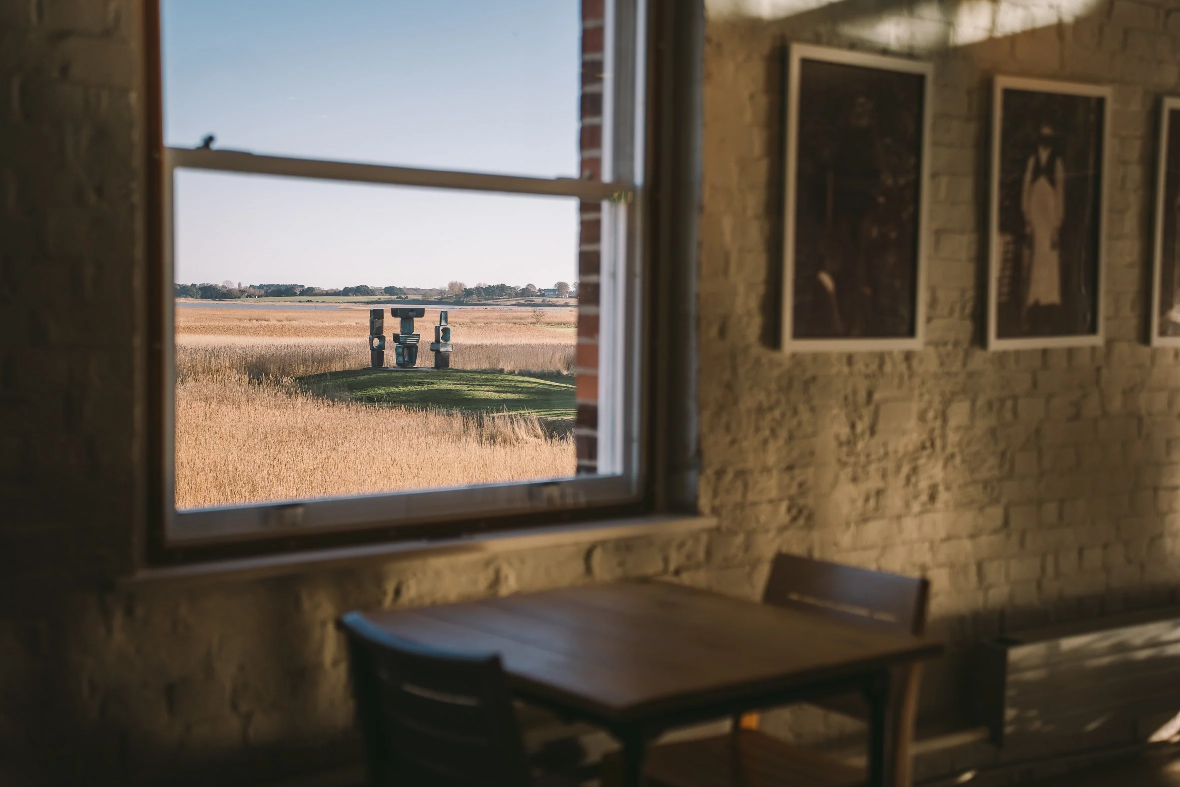 View of an open window with a brick wall inside a room, showing a scenic landscape outside with tall grasses and sculptures in the distance.
