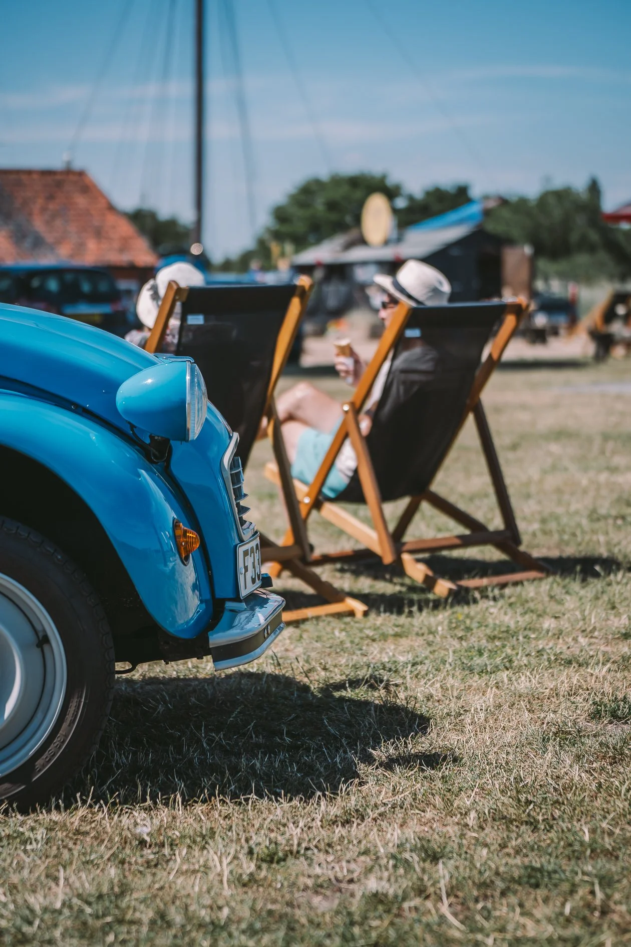 A close-up of a vintage blue car in the foreground with a woman relaxing on a deck chair in the background, at an outdoor setting on a sunny day.