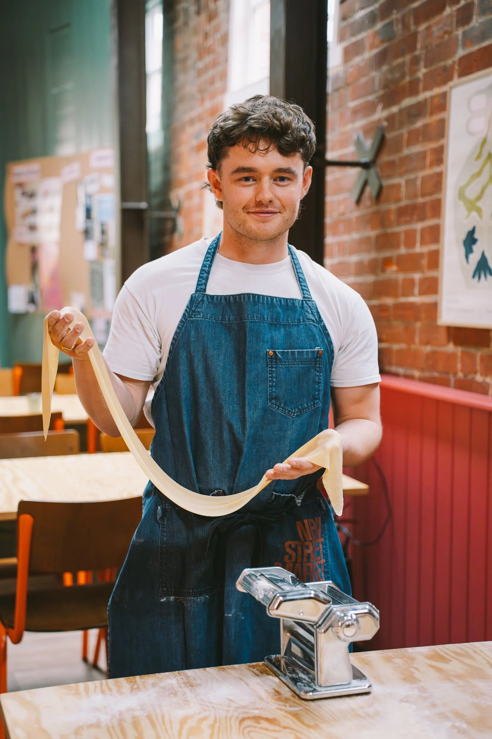 A young man with dark, curly hair wearing a white t-shirt and a blue apron, making pasta in a kitchen with wooden tables and brick walls.