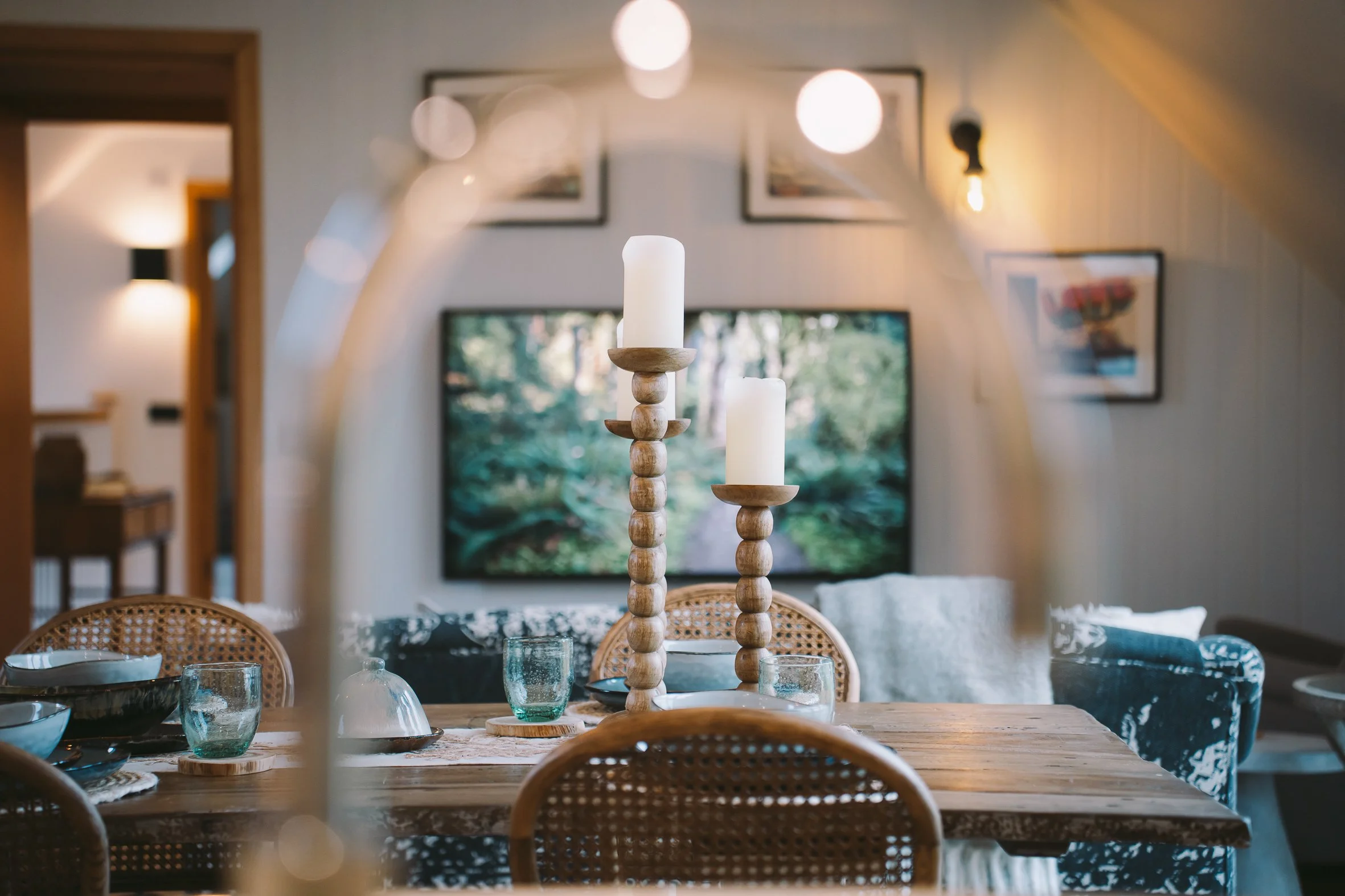 A dining table set with glassware and surrounded by wicker chairs, with a wooden candle holder centerpiece on a tablecloth in a cozy living space with wall art and warm lighting.