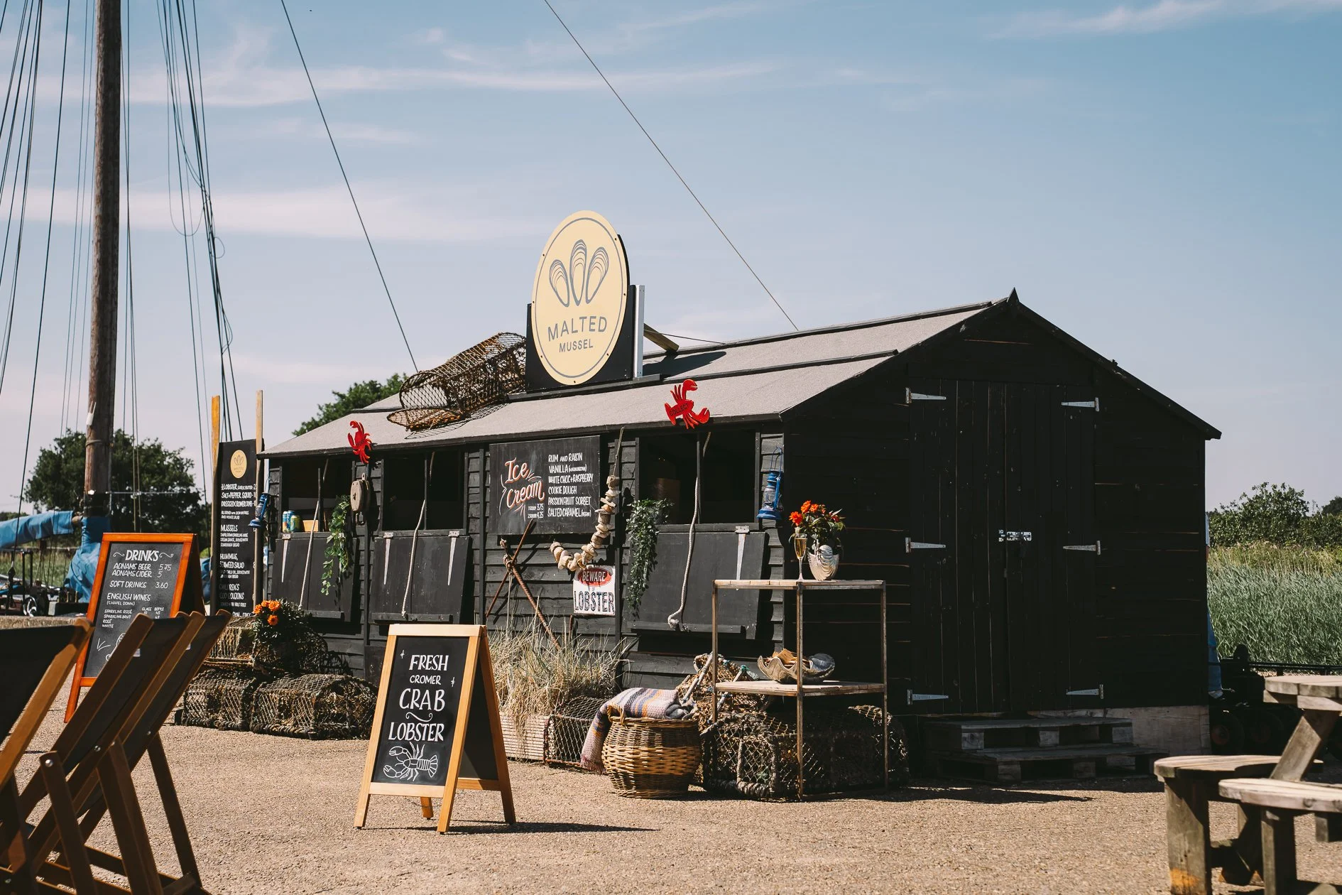 A small black wooden building with a sign that says 'Malted Mussel' on the roof, decorated with crew and nautical items, and surrounded by outdoor seating, menu boards, and baskets on a sunny day with a clear blue sky.