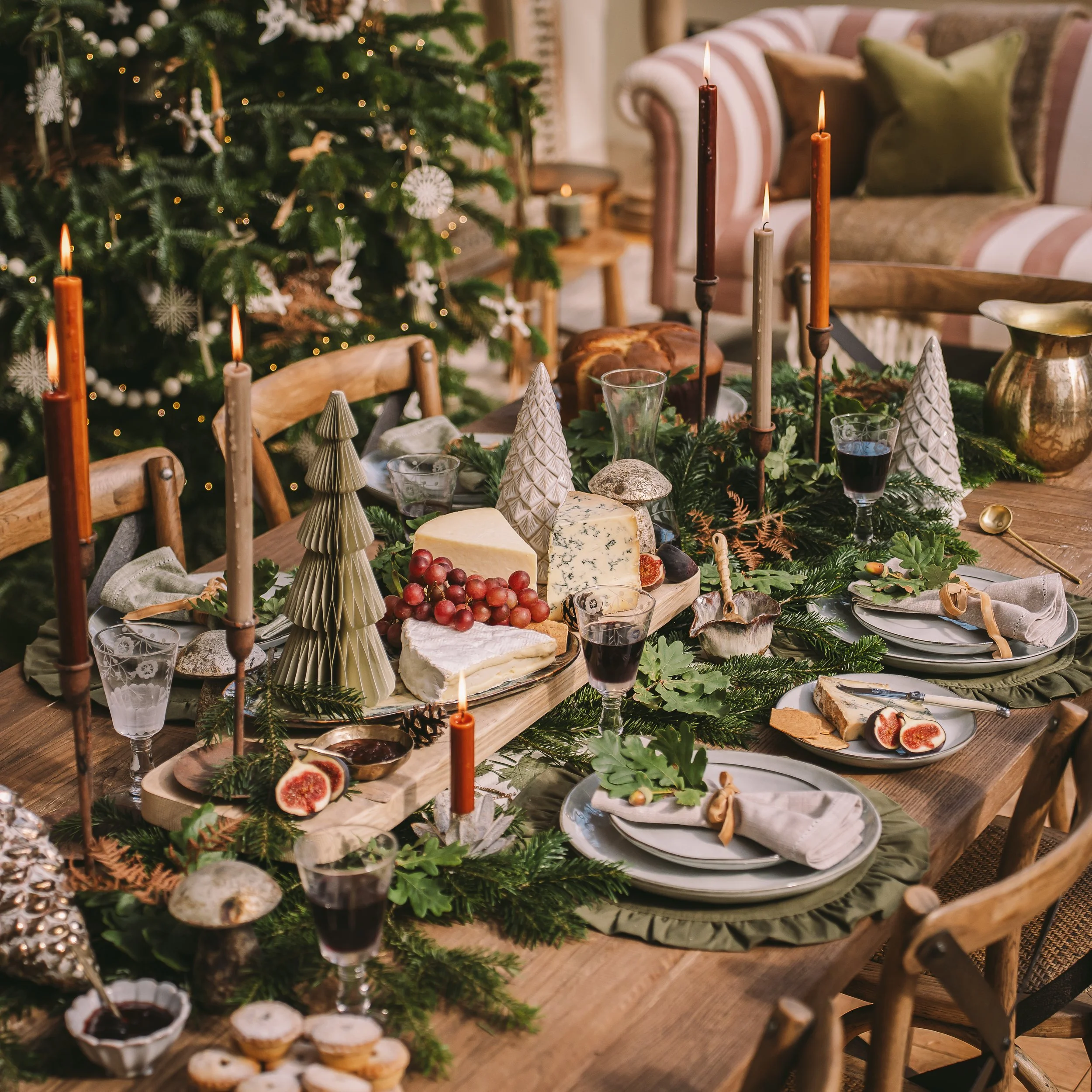 A festive holiday table decorated with greenery, candles, cheeses, figs, grapes, and wine glasses, with a decorated Christmas tree and a cozy living room in the background.