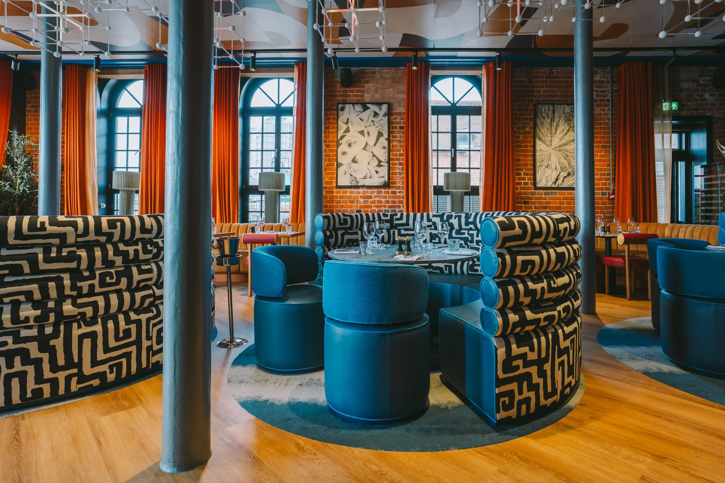 Interior of a modern restaurant or lounge with large windows, orange curtains, geometric patterned seating, and abstract wall art.