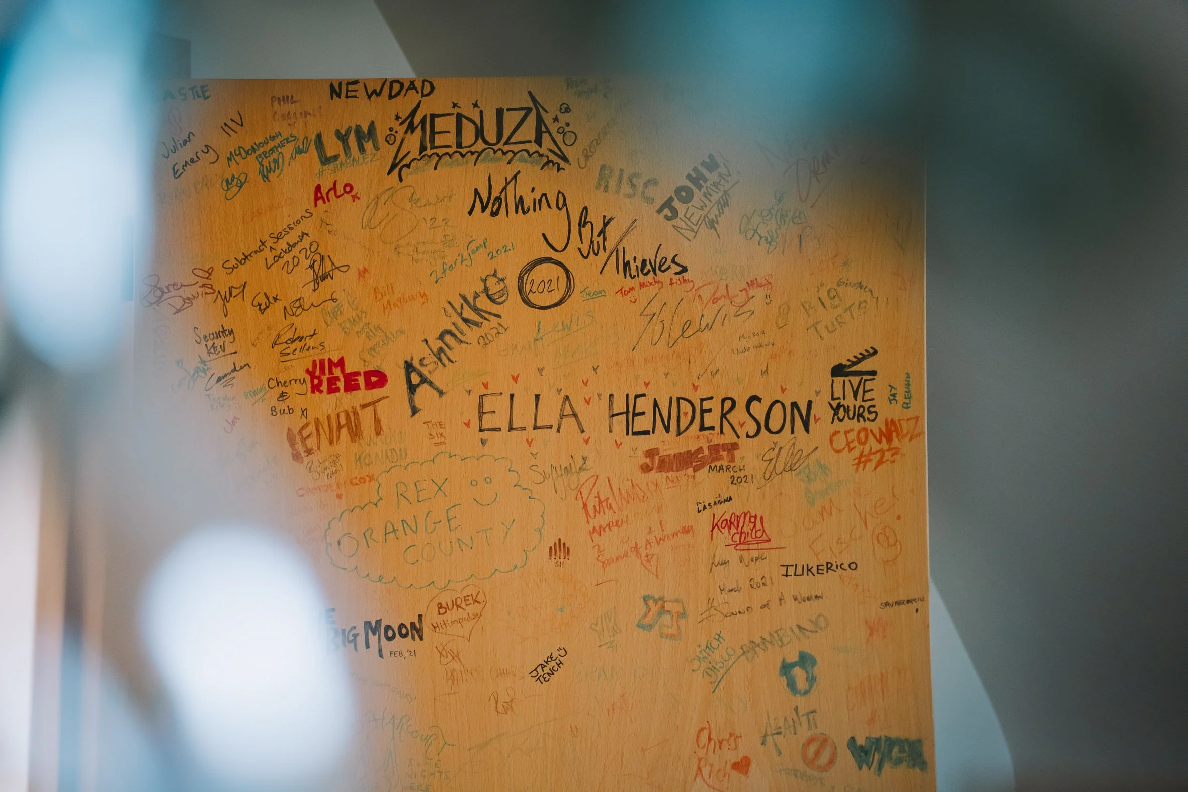 A wall covered with various handwritten signatures and messages in different colors, including names like Ella Henderson, Jim Reed, Lyin Meduza, and others, with some drawings and doodles.
