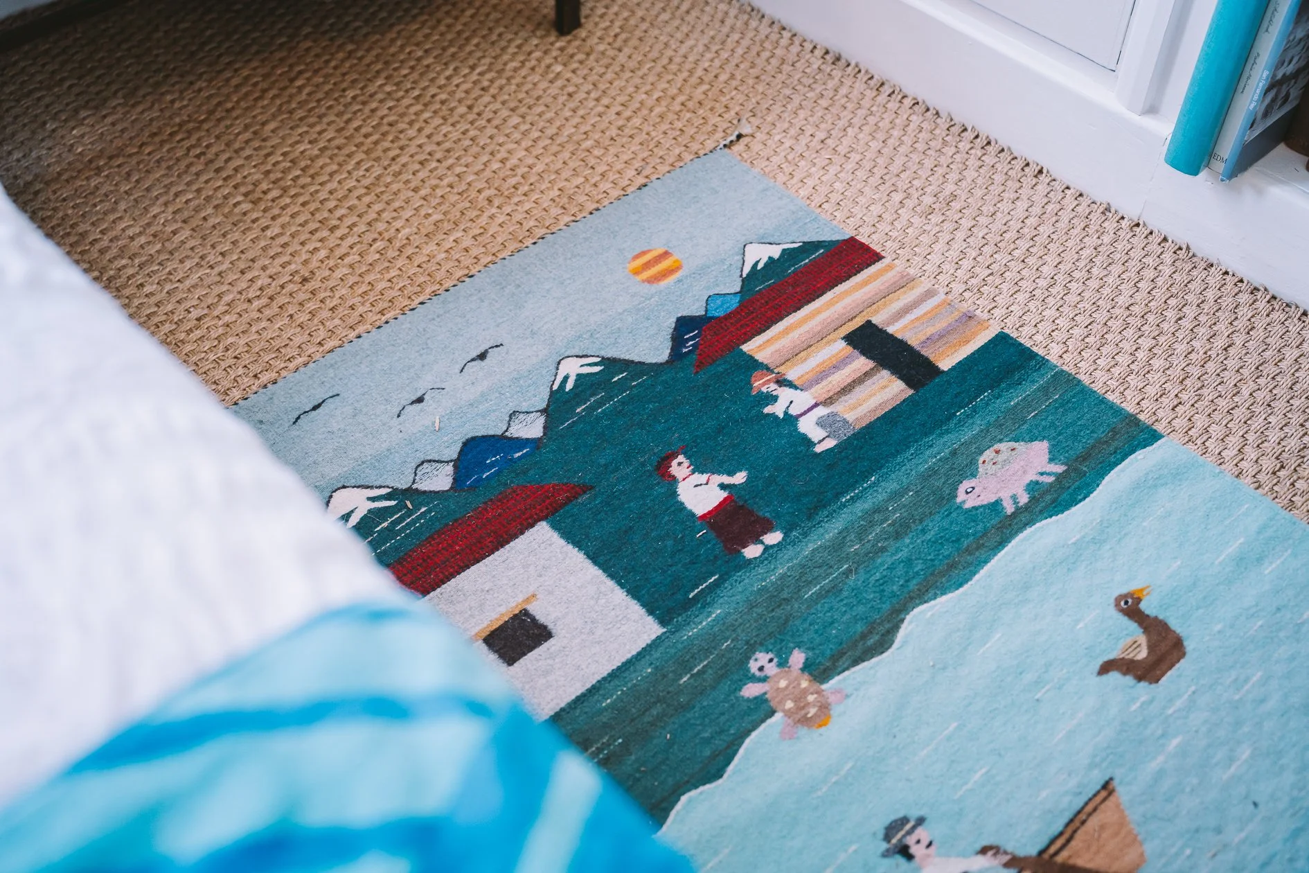 Part of a colorful illustrated rug depicting mountains, a sun, animals, and a child, on a beige carpet next to a white wall with a white shelf containing books.