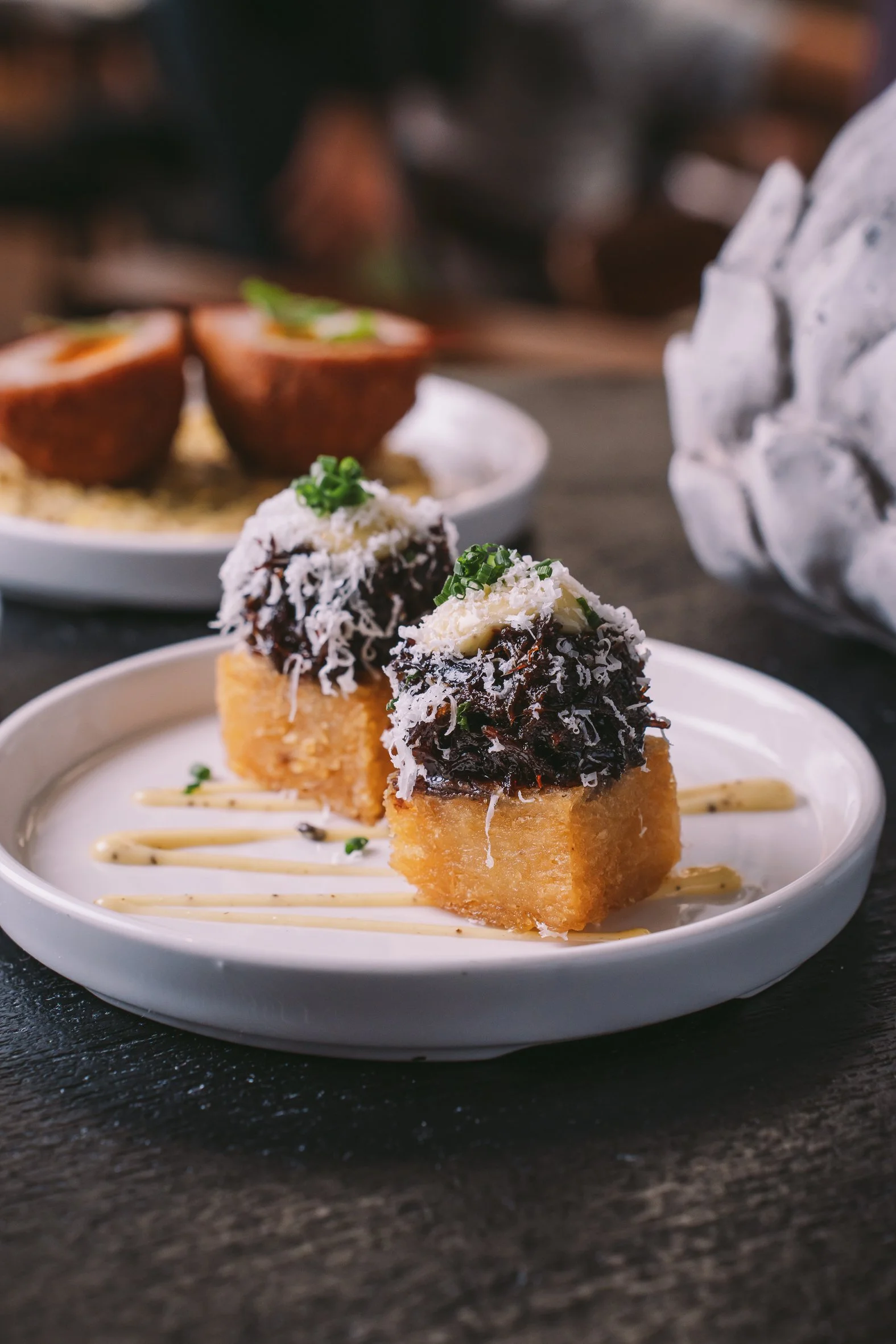Close-up of two savory appetizers on a white plate, topped with shredded cheese and chopped green onions, served with a drizzle of sauce on a dark surface.