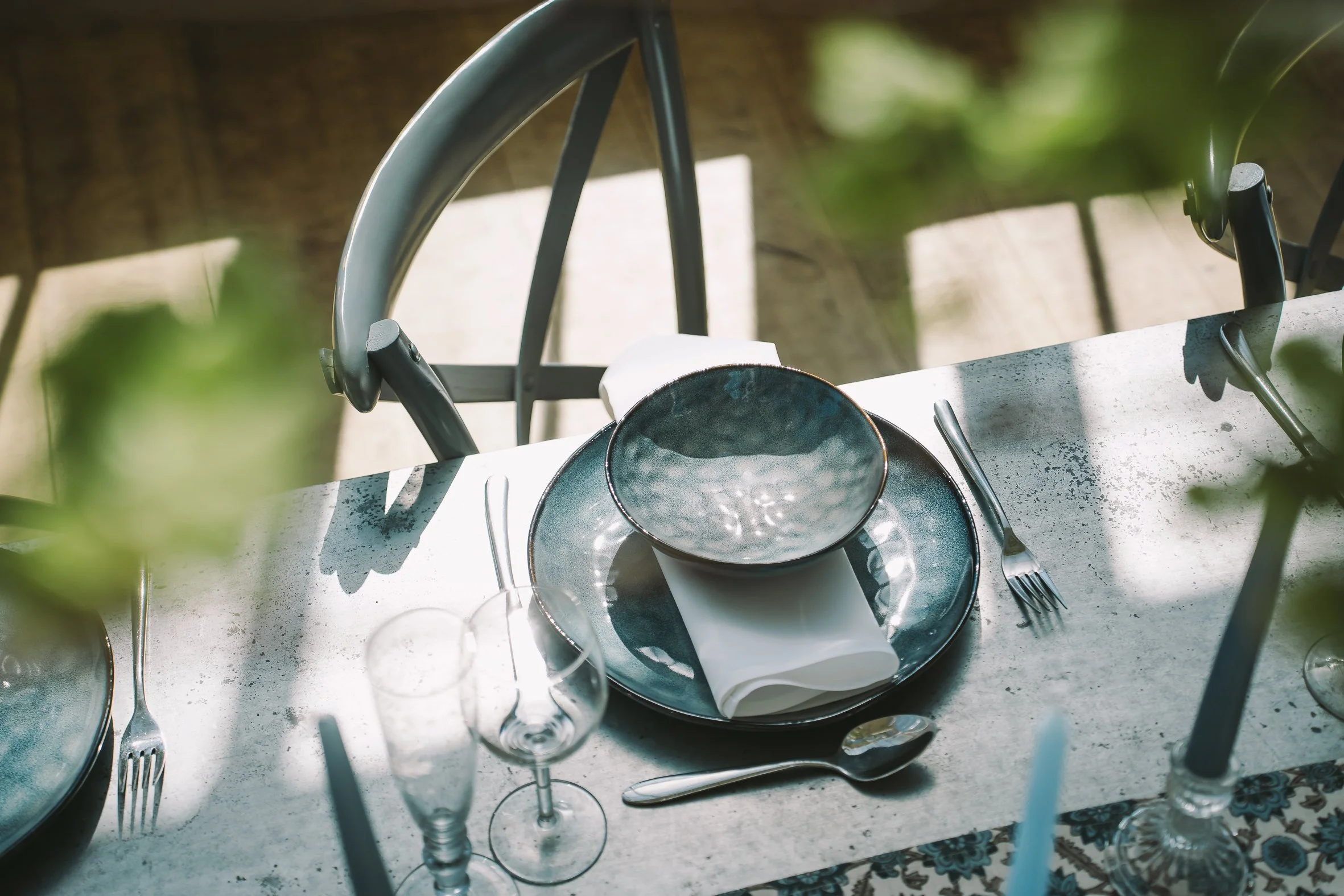 A table set for a meal outdoors with a cup and saucer, wine glass, fork, knife, and spoon, with blurred greenery in the background.