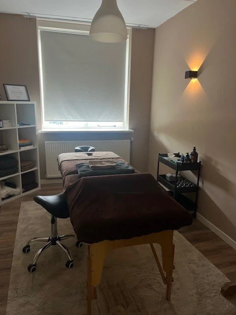 Een massagekamer met een massagetafel, een bureaustoel, een boekenkast, en een raam met een gesloten rolgordijn.