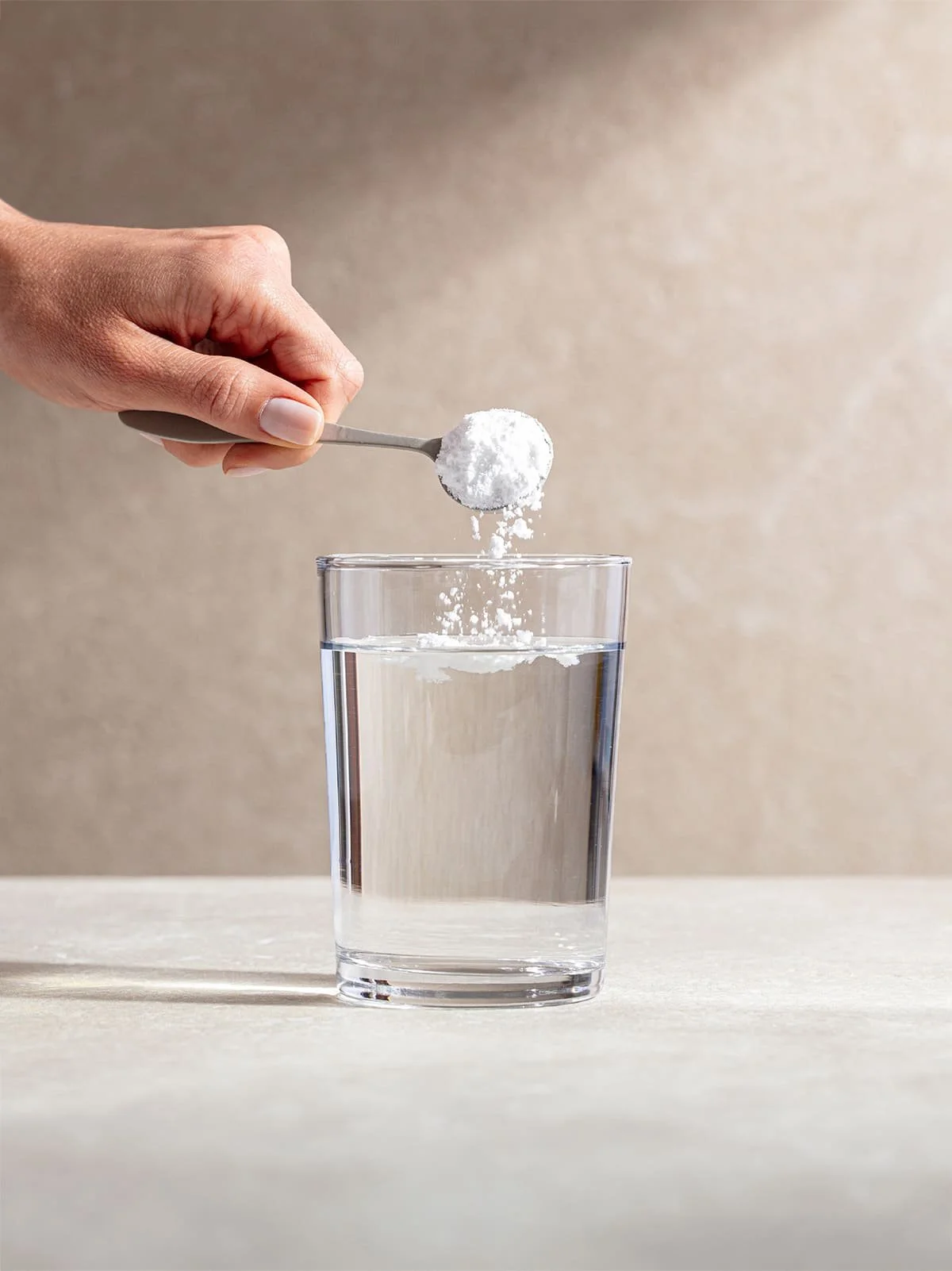 Een hand die een witte poeder, mogelijk bicarbonaat, van een lepeltje in een glas water strooit.