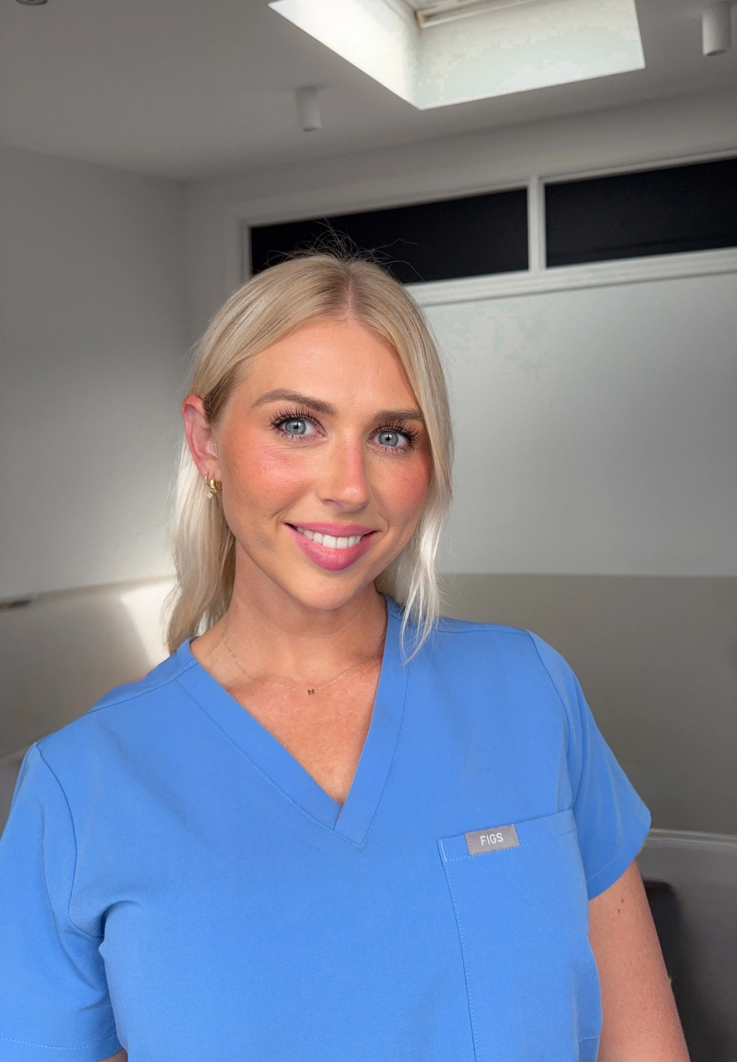 Close-up of a woman with blonde hair and blue eyes wearing blue medical scrubs with a name tag that reads 'FIGS' in a medical setting.