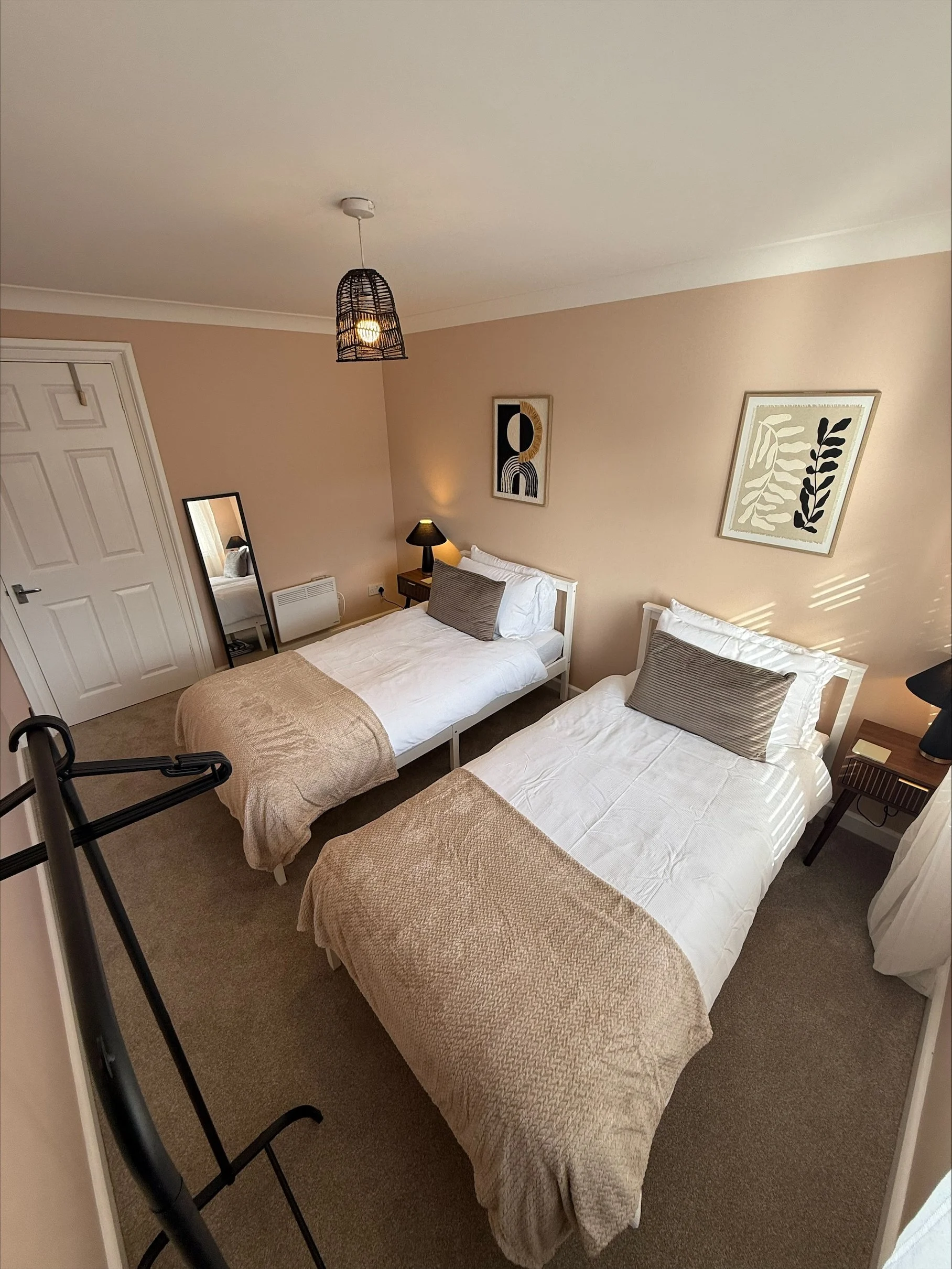 Cozy bedroom with two twin beds, beige textured blankets, woven pendant light, bedside tables, and botanical wall art