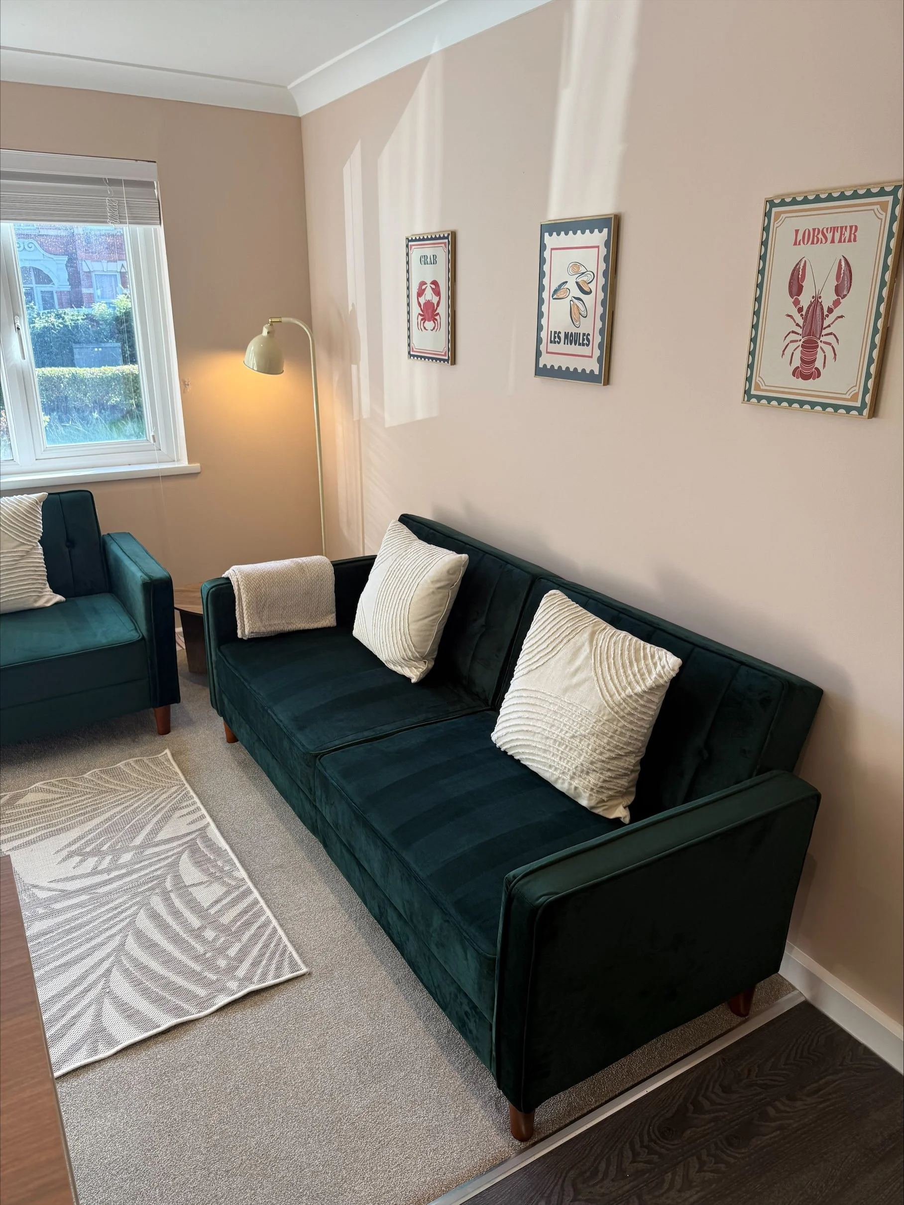 Coastal-themed living room with dark teal sofa and armchair, cream pillows, floor lamp, window, patterned rug, and framed crab, mussel, and lobster artwork