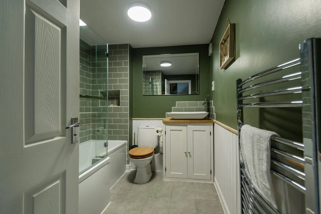 Modern green and white bathroom with bathtub, glass shower screen, toilet, vanity, mirror, and chrome towel radiator