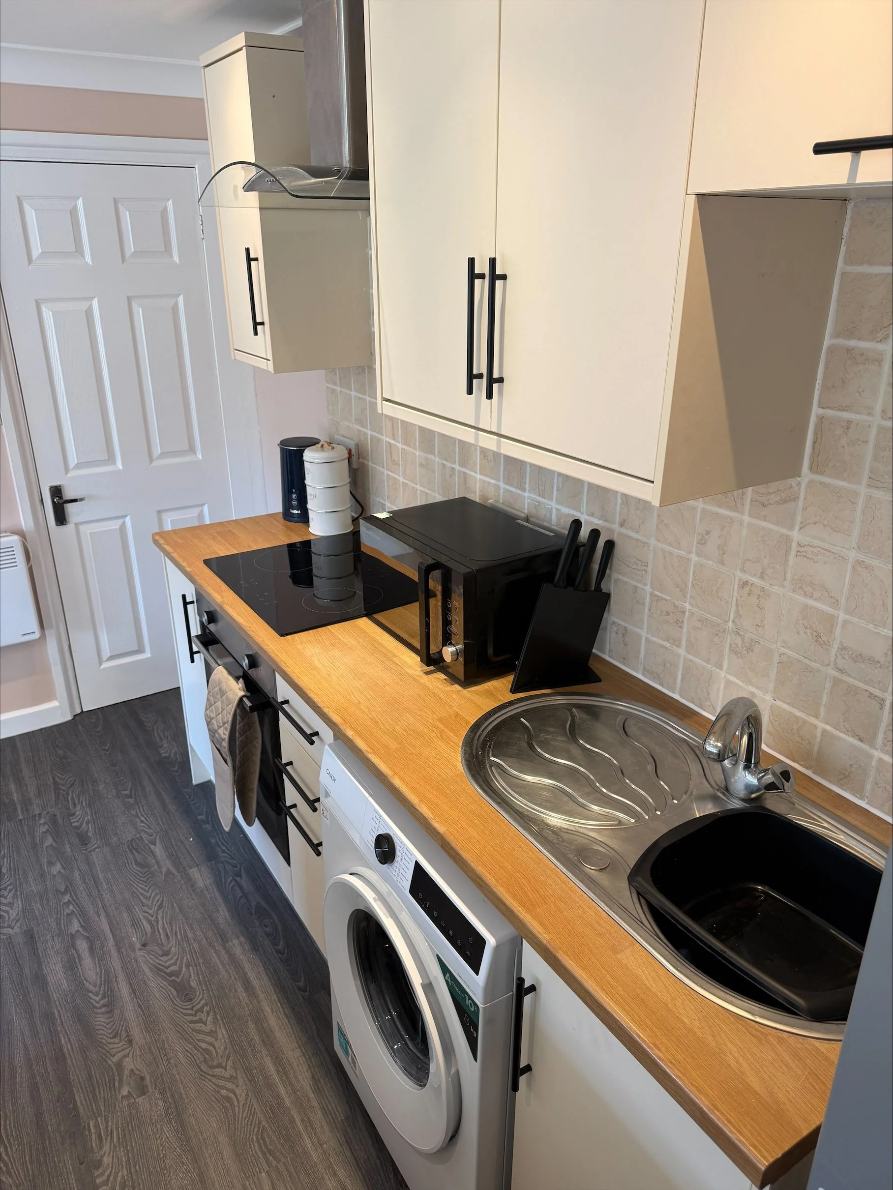 Compact kitchen with cream cabinets, wood countertop, black induction cooktop, appliances, double sink, and tiled backsplash