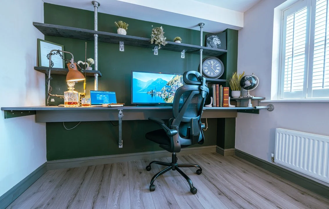 Modern home office with a wall-mounted desk, ergonomic chair, computer monitors, industrial pipe shelving, and window shutters