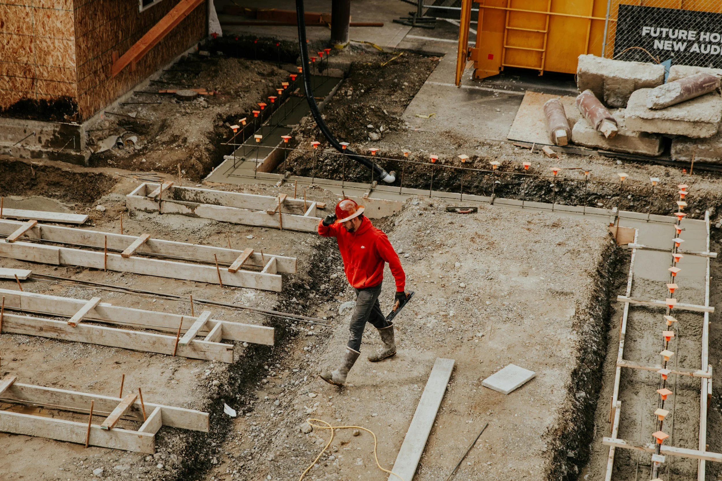 Construction workers at a building site, preparing the foundation with wooden forms and concrete, surrounded by dirt, construction materials, and tools.