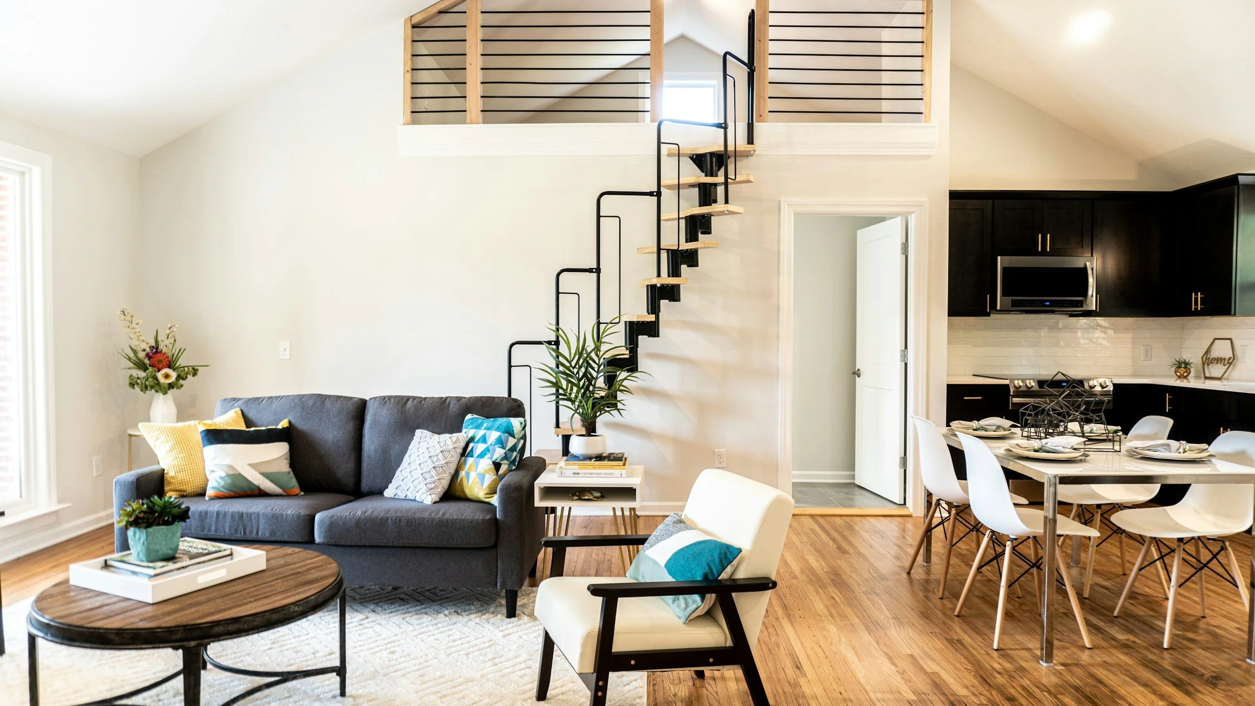 Open-concept living room and dining area in a modern home with a loft, black staircase, white walls, and wooden flooring. The living room has a gray sofa with colorful pillows, a small side table, and a coffee table with a potted plant. The dining area features a glass table with white chairs.