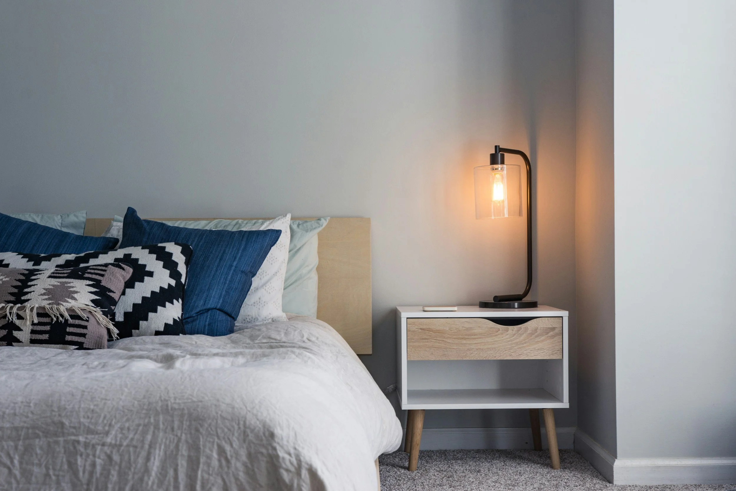 A neatly made bed with white and navy blue bedding and patterned pillows, a white nightstand with a wooden drawer, and a modern black floor lamp with a glass cover on a gray wall in a bedroom.