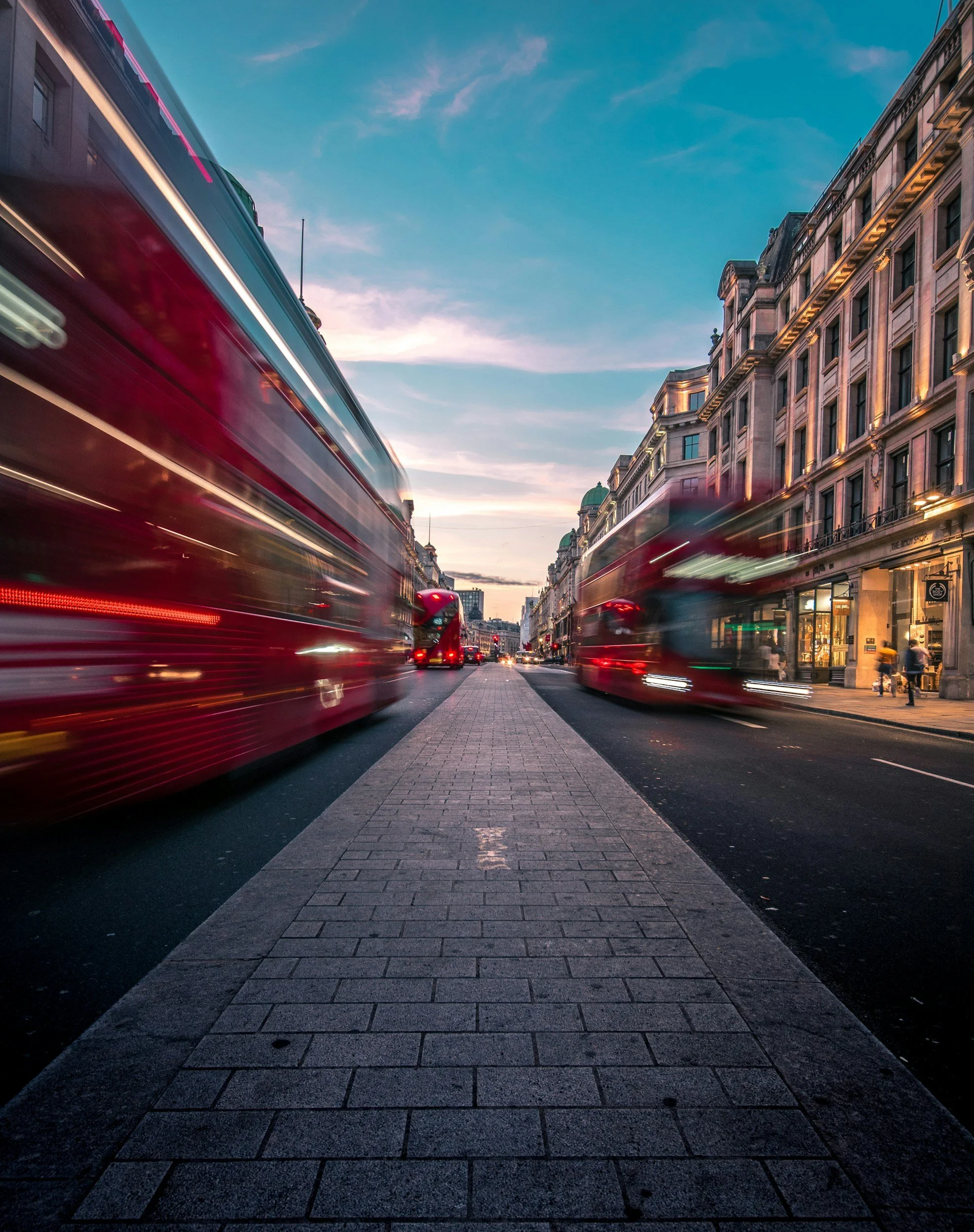 City street at dusk with blurred red double-decker buses and historic buildings on either side.