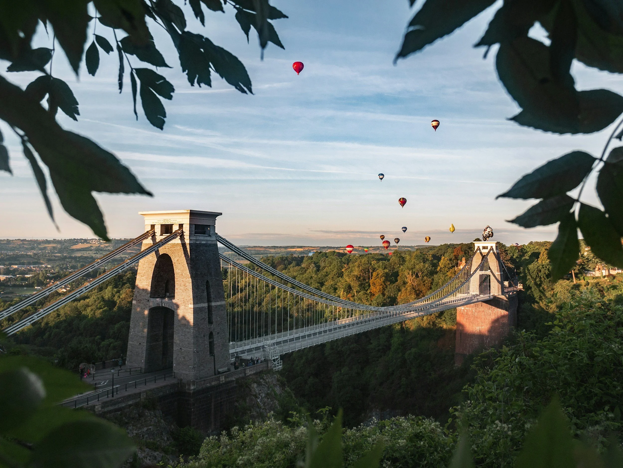 Several hot air balloons floating in the sky over a bridge, framed by leafy branches in the foreground, with a landscape and buildings below.