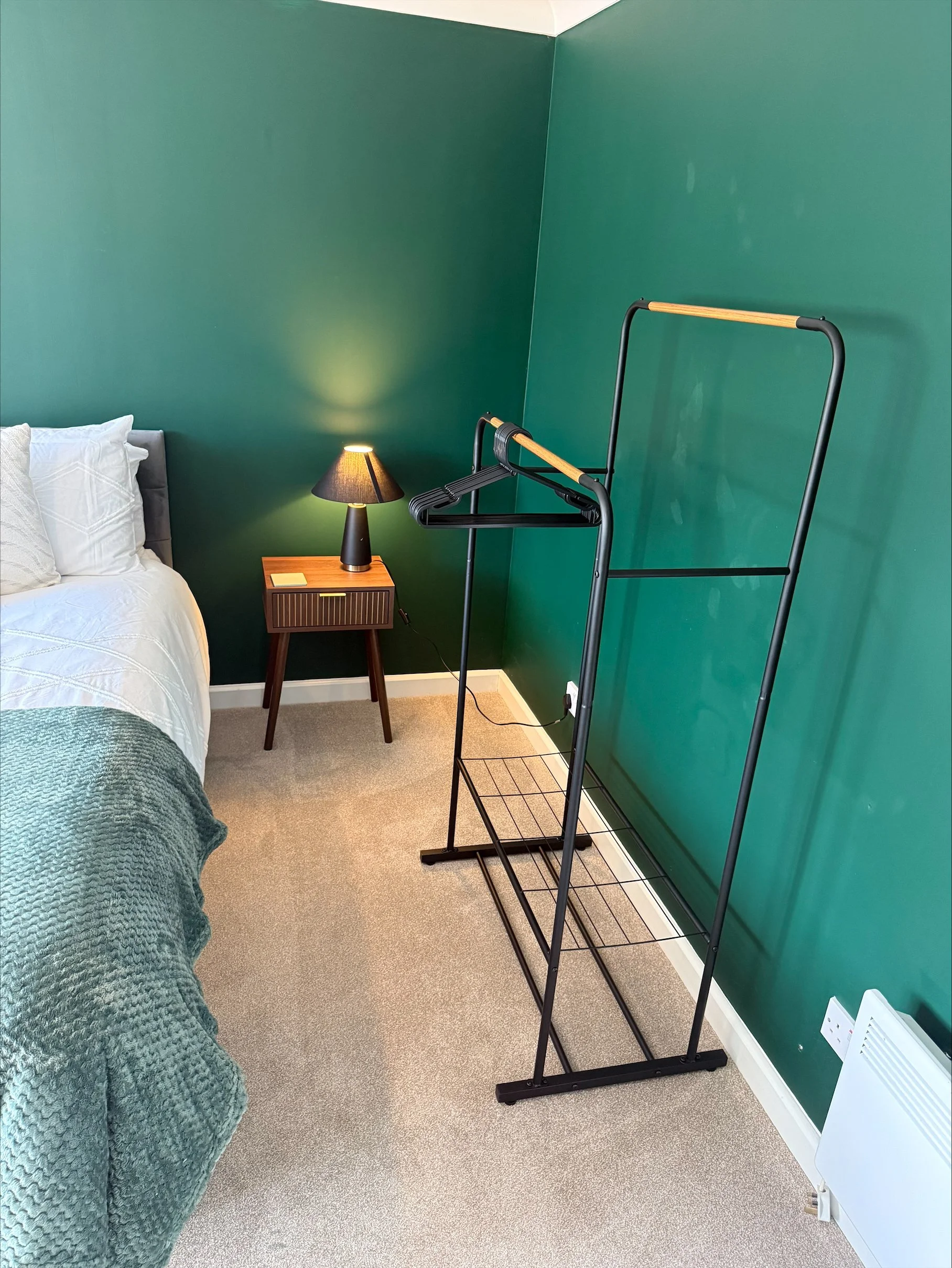 Cozy bedroom with a white bed, green textured blanket, bedside table and lamp, and a black wooden clothes rack against dark teal walls.