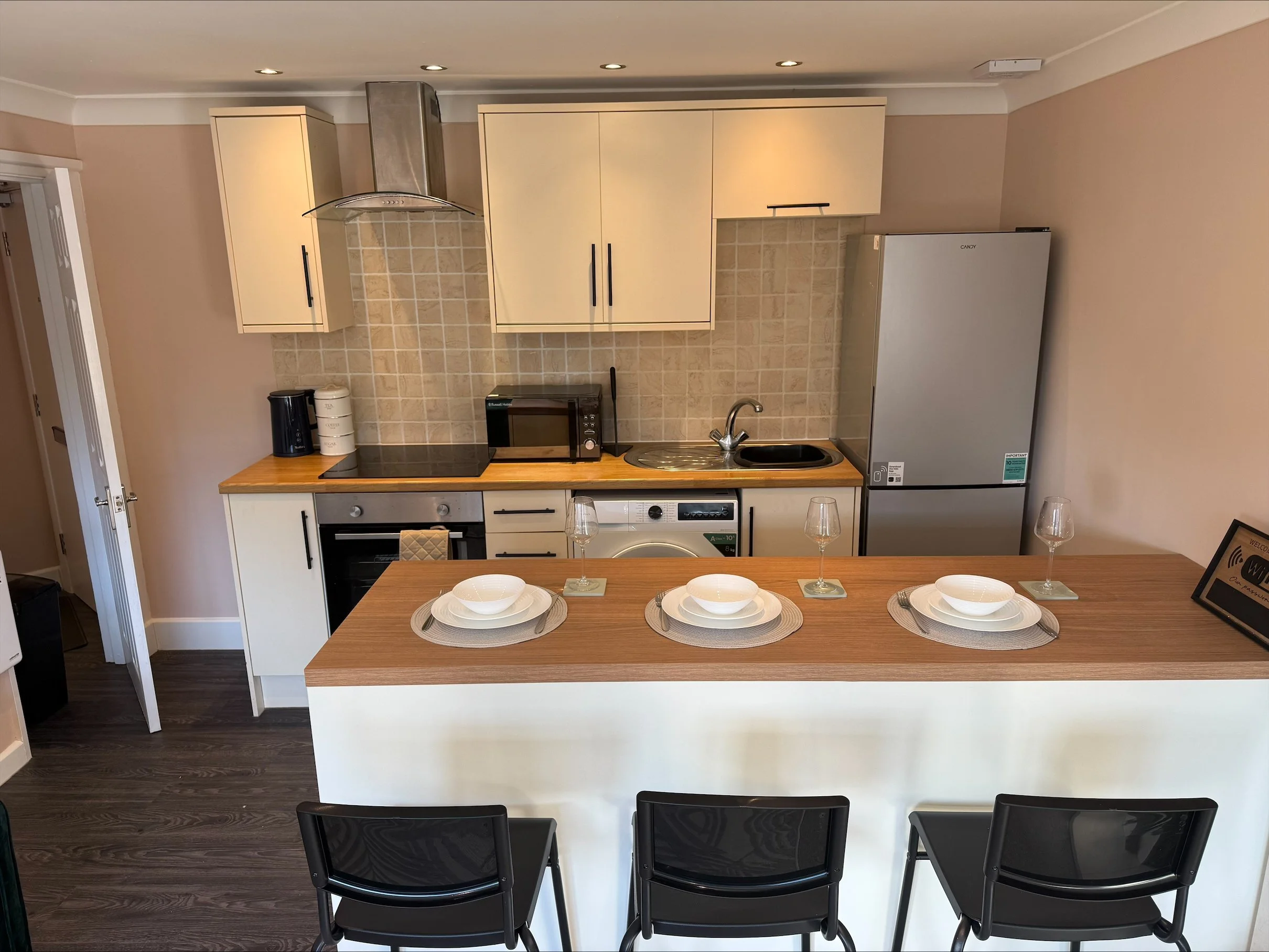 Modern kitchen with cream cabinets, tiled backsplash, appliances, breakfast bar, place settings, and three black bar stools