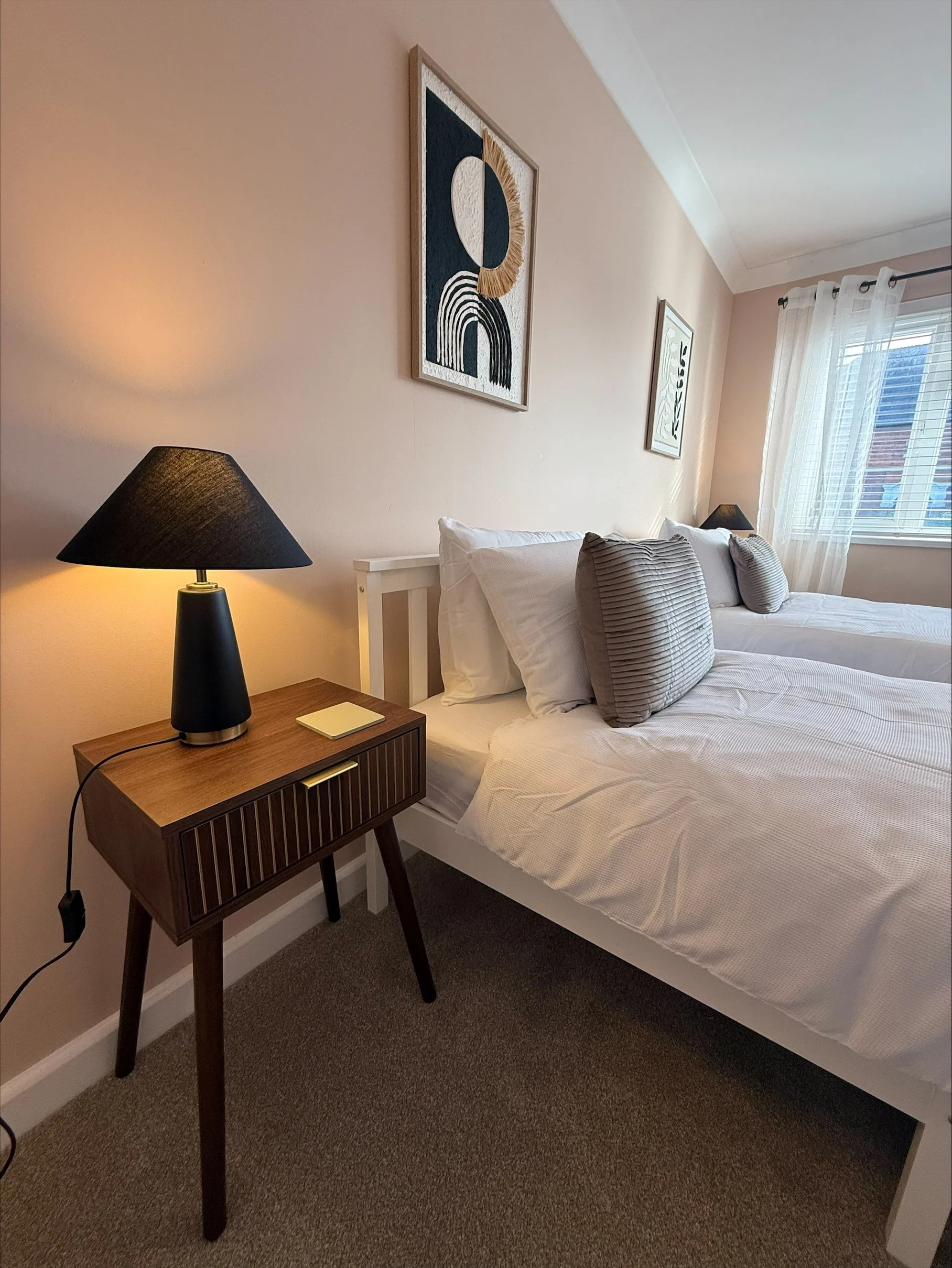Cozy modern bedroom with a neatly made white bed, black bedside lamp, wood nightstand, abstract wall art, and window with sheer curtains.