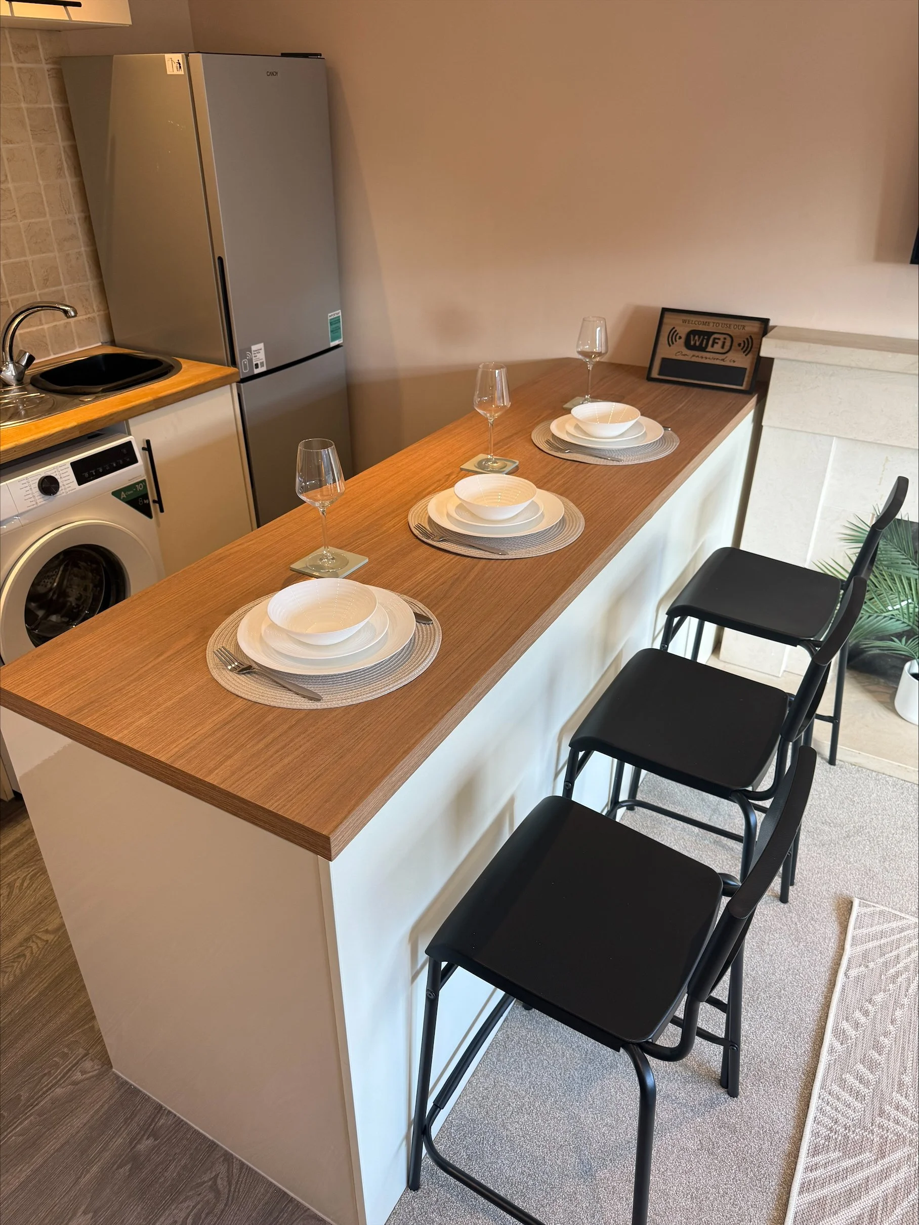 Modern apartment kitchen with a breakfast bar set for three, black bar stools, refrigerator, sink, washer, and Wi-Fi sign