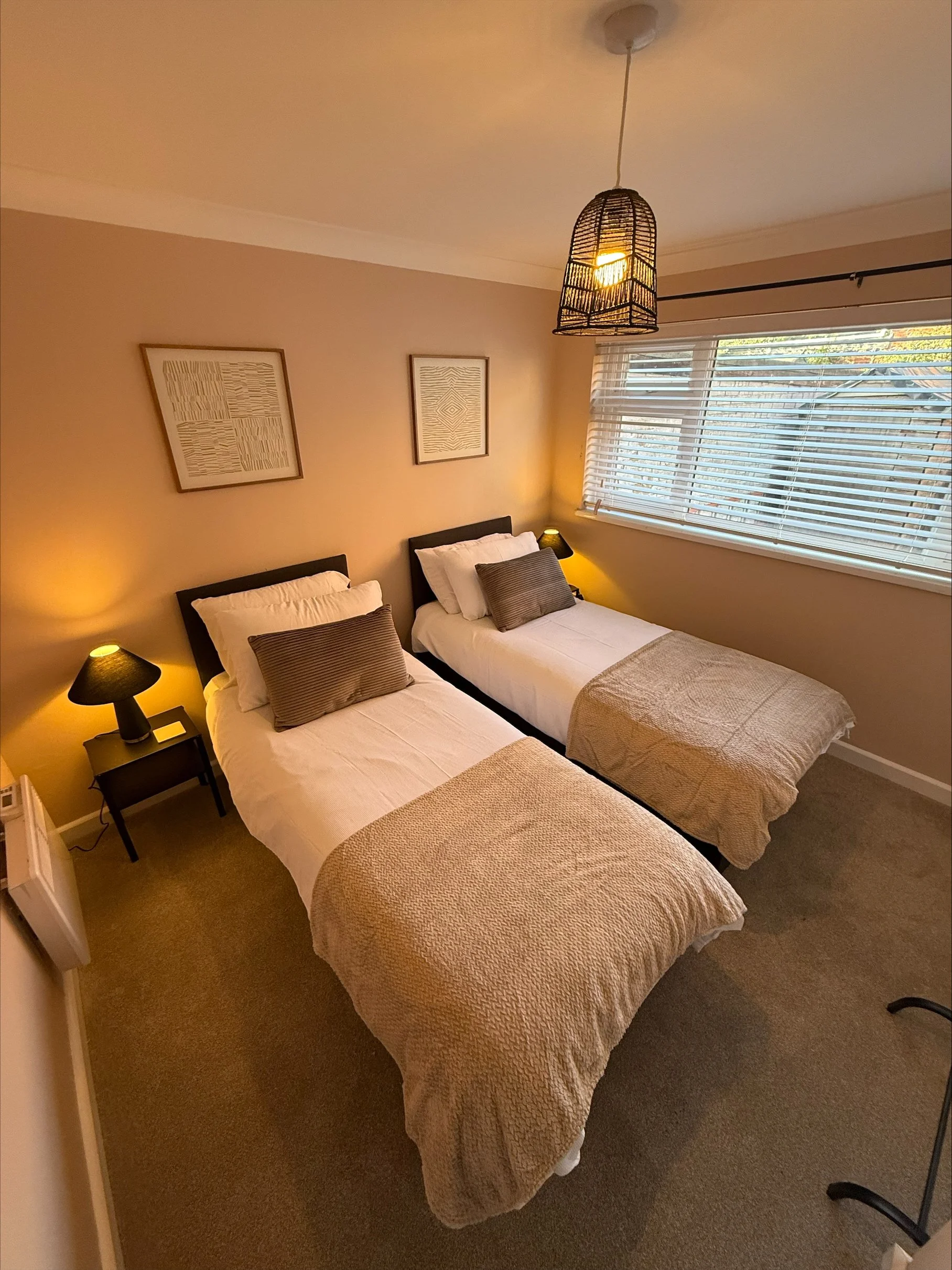 Cozy bedroom with two neatly made twin beds, bedside lamps, framed artwork, woven pendant light, and window blinds