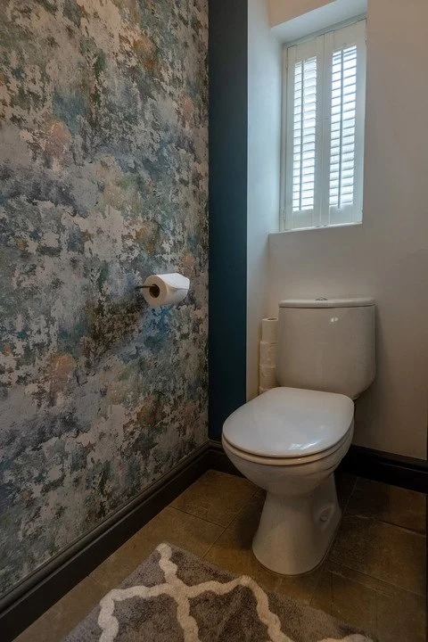 Small bathroom with a white toilet, patterned wallpaper, plantation-shuttered window, and area rug