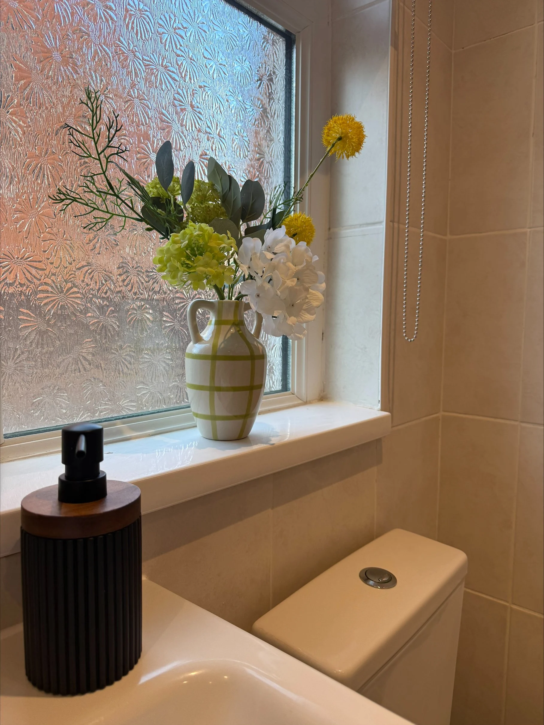 Bathroom vanity with a green-and-white floral vase beside a frosted floral window, soap dispenser, and toilet