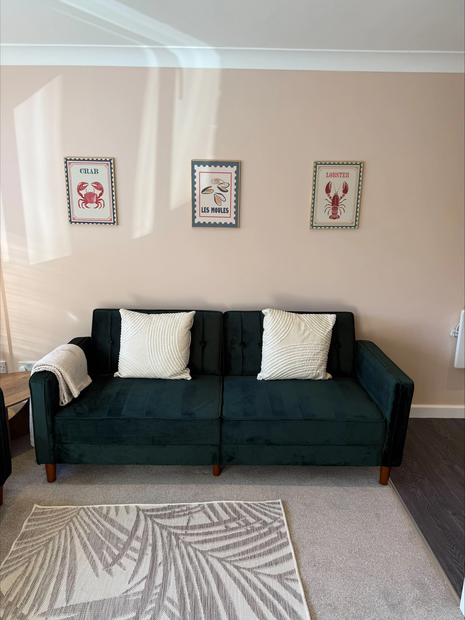 Dark green velvet sofa with white pillows and throw blanket beneath framed crab, mussel, and lobster artwork in a sunlit living room