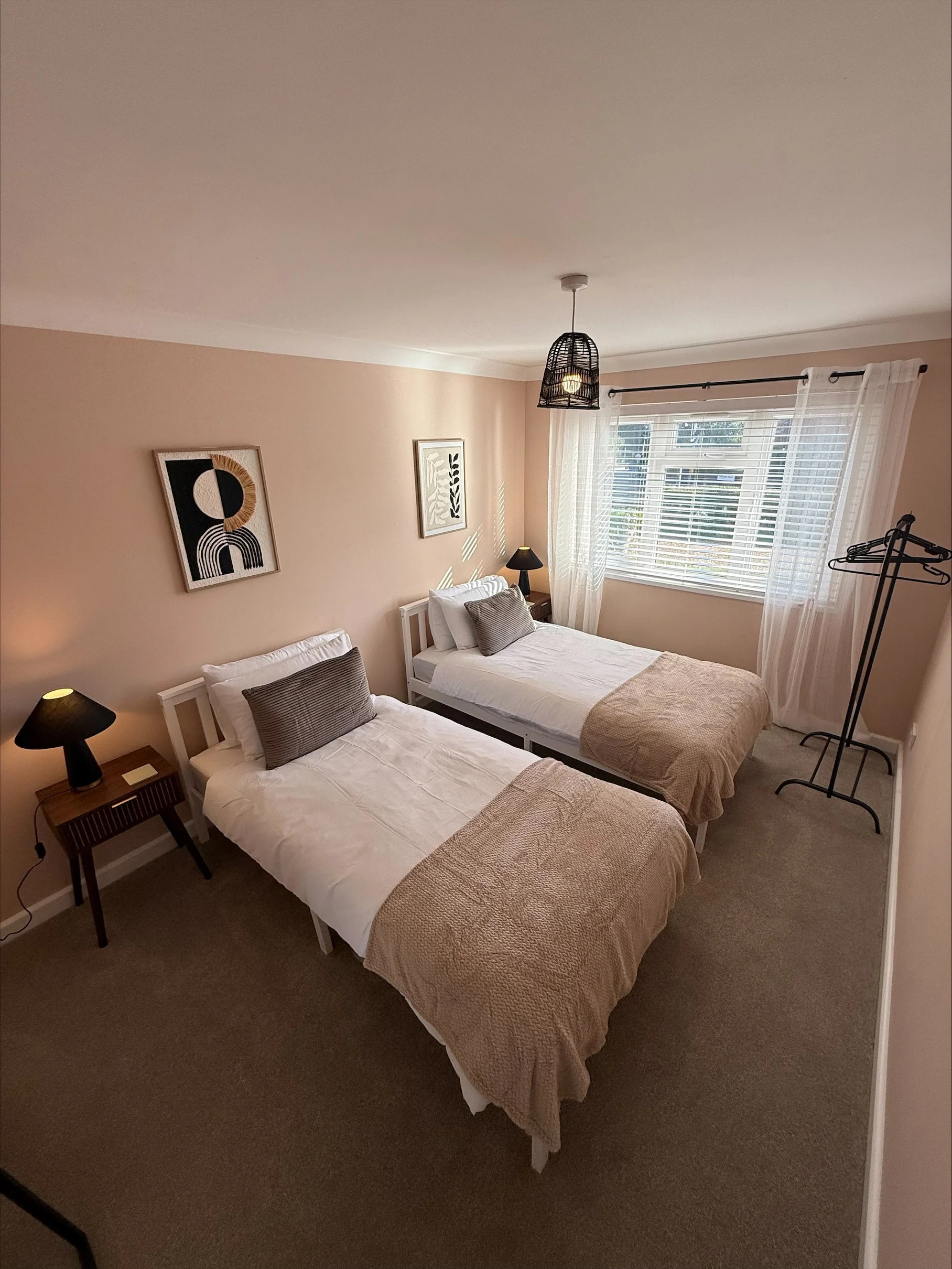 Bright bedroom with two neatly made twin beds, beige throws, bedside lamps, wall art, a woven pendant light, and a sunlit window with sheer curtains.