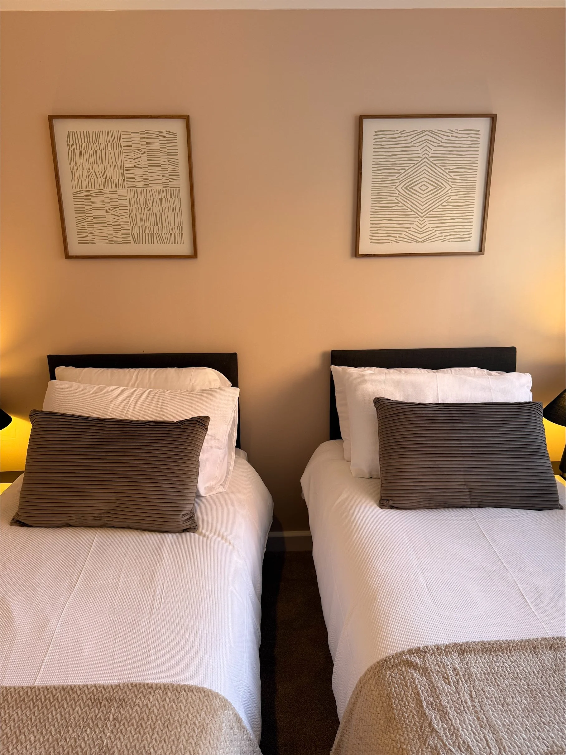 Cozy bedroom with two twin beds, white bedding, gray pillows, warm bedside lighting, and abstract framed artwork