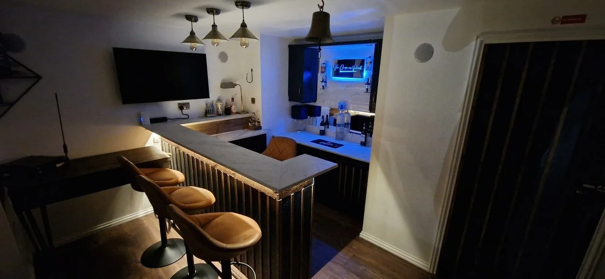 Dimly lit home bar with three brown stools, marble counter, liquor bottles, hanging pendant lights, and wall-mounted TV