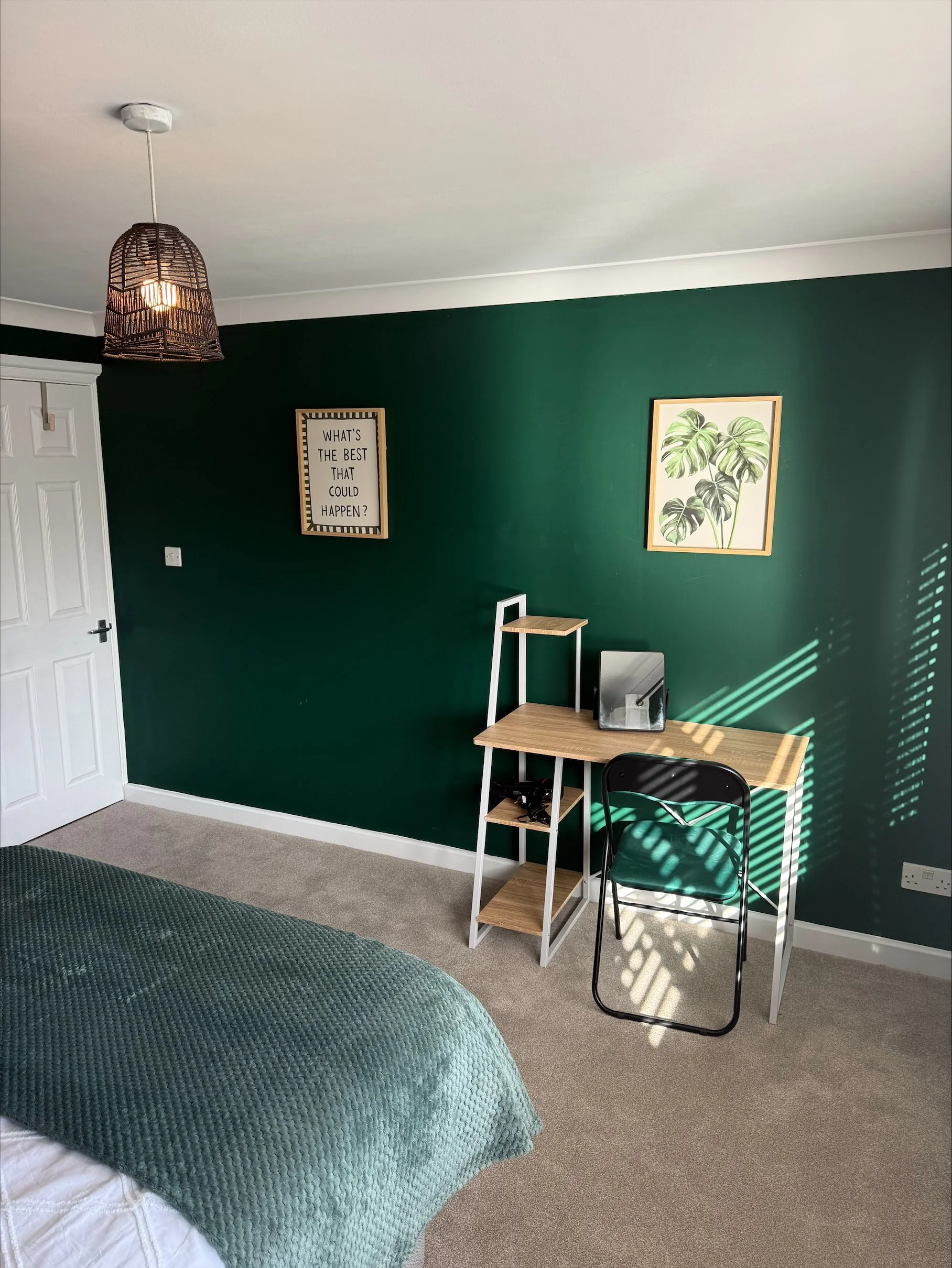 Sunlit bedroom with a dark green accent wall, wood desk, black folding chair, framed leaf artwork, motivational sign, and woven pendant light.