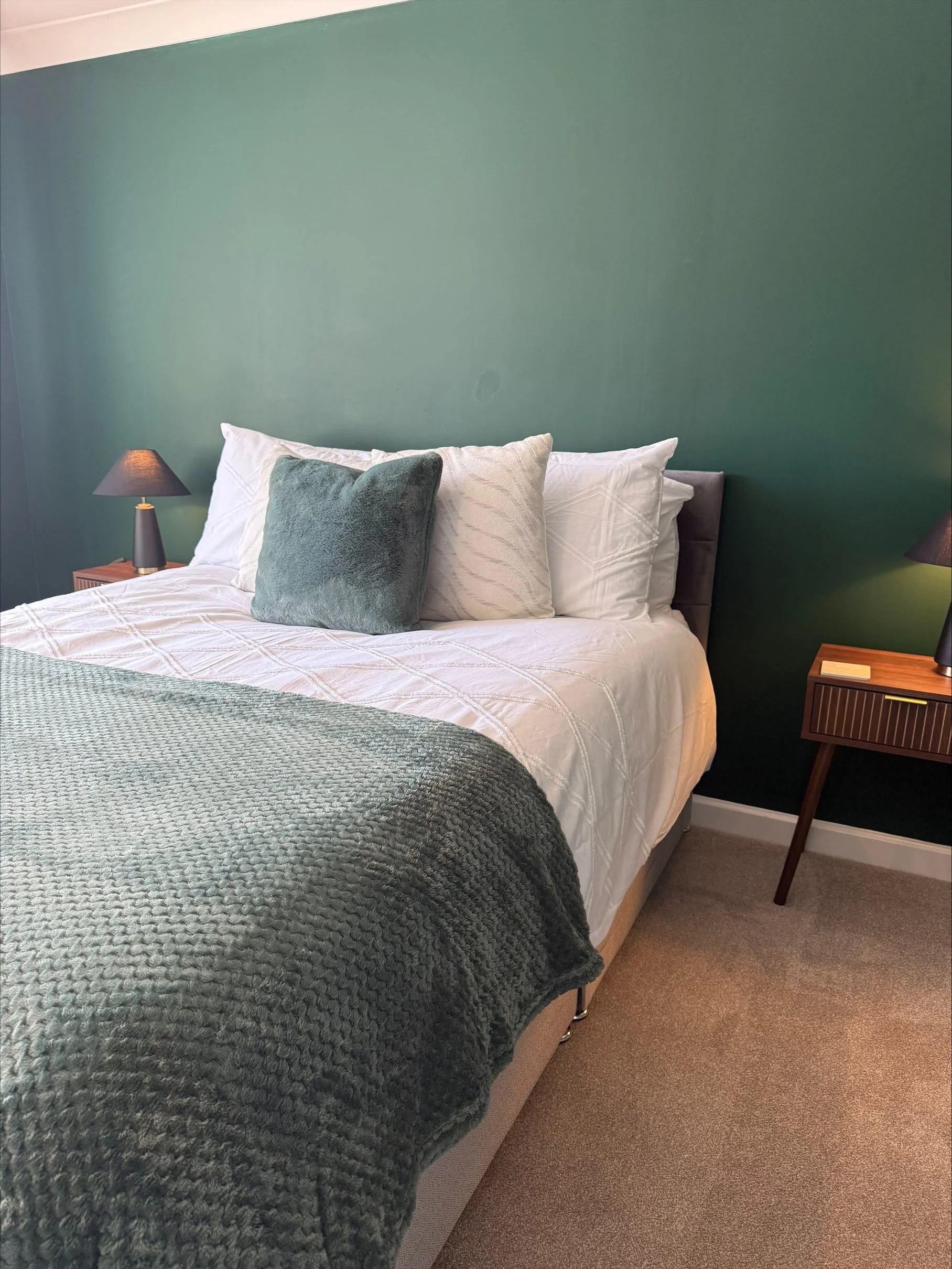Cozy bedroom with a neatly made bed, white bedding, teal throw blanket and pillows, dark green accent wall, and matching bedside lamps and nightstands.