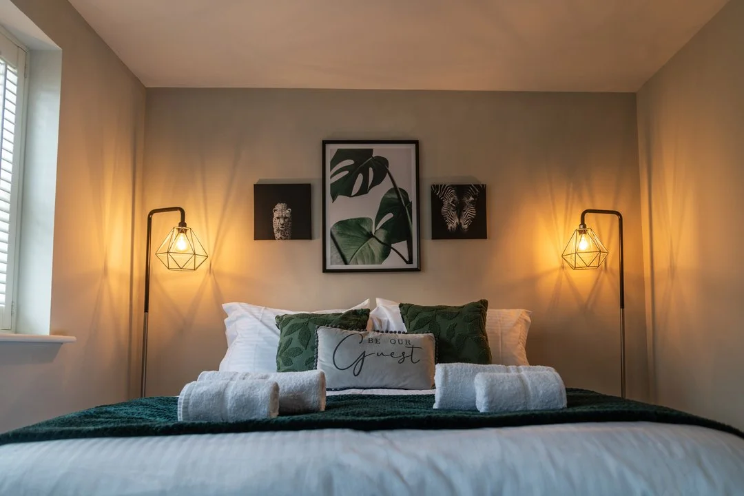 Cozy guest bedroom with a neatly made bed, green leaf-patterned pillows, “Be Our Guest” accent pillow, warm bedside lamps, and botanical wall art.