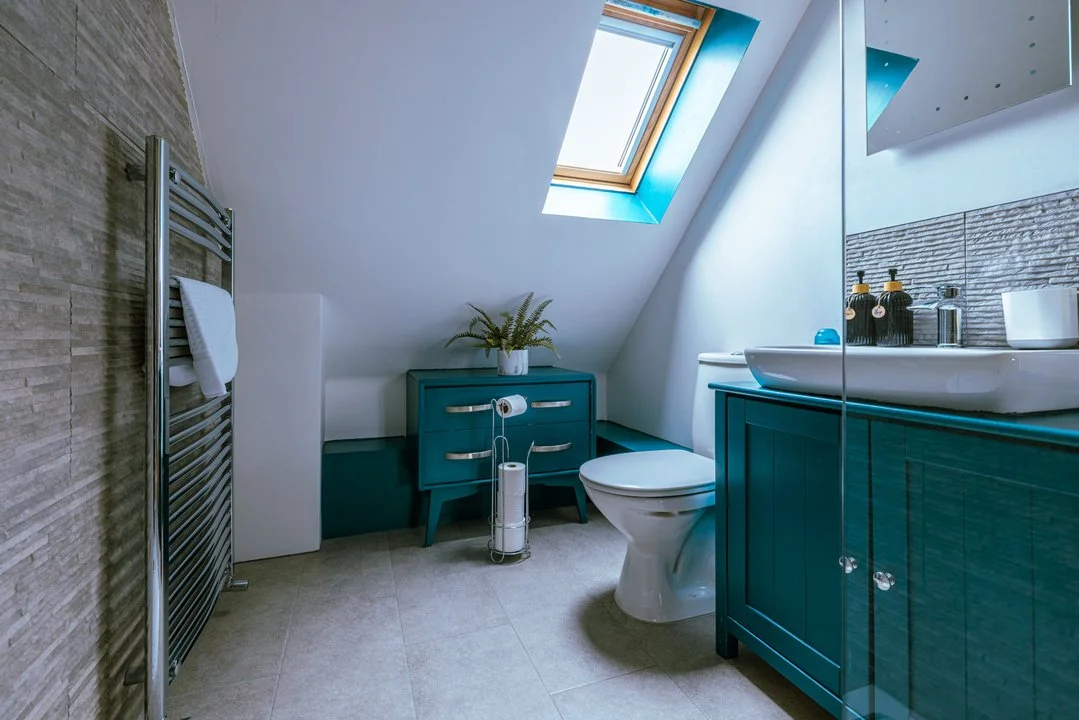 Modern attic bathroom with skylight, teal vanity, white toilet, heated towel rack, and textured stone walls