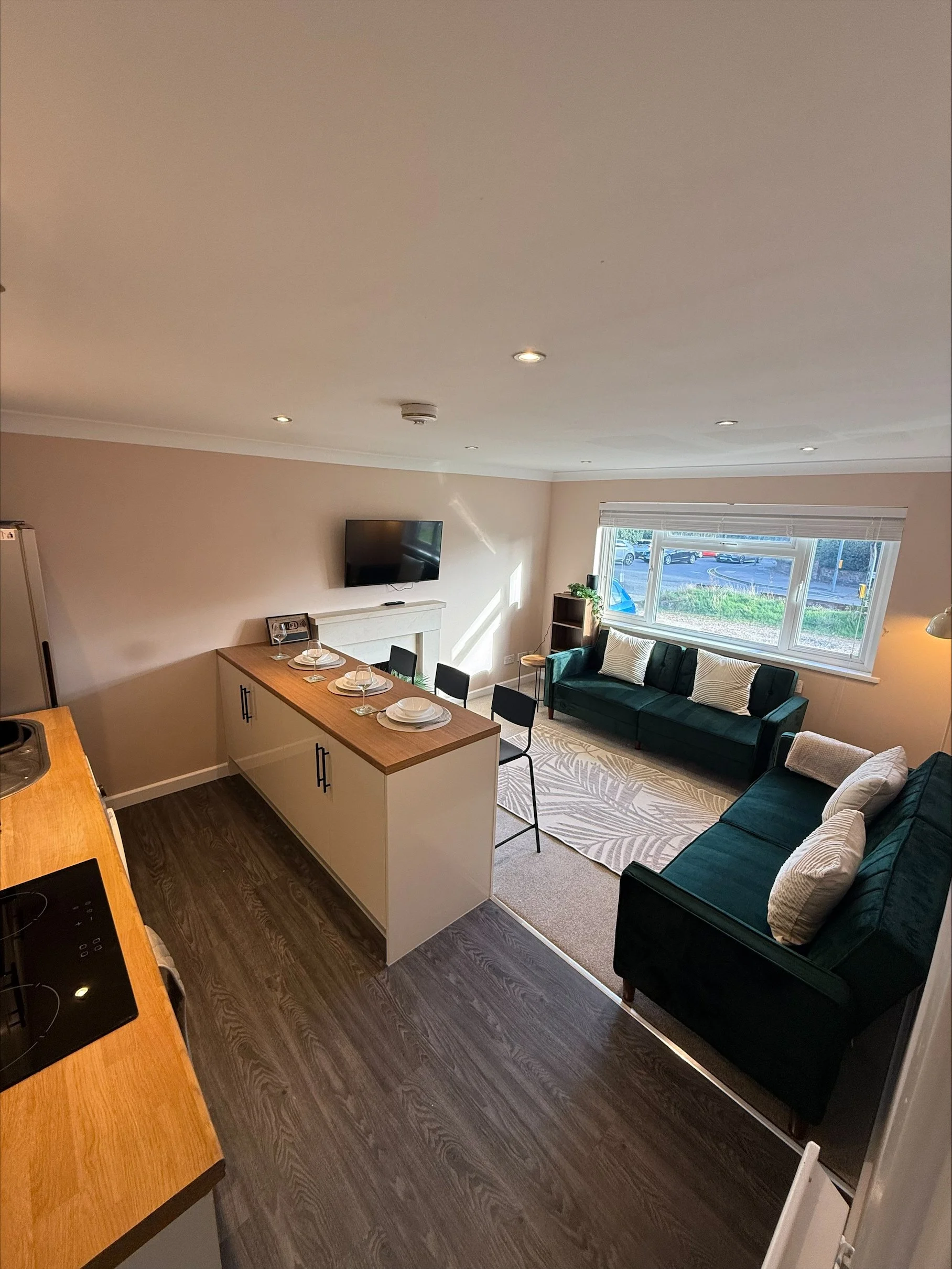 Modern open-plan kitchen and living room with island dining table, wall-mounted TV, fireplace, teal sofas, and large window.