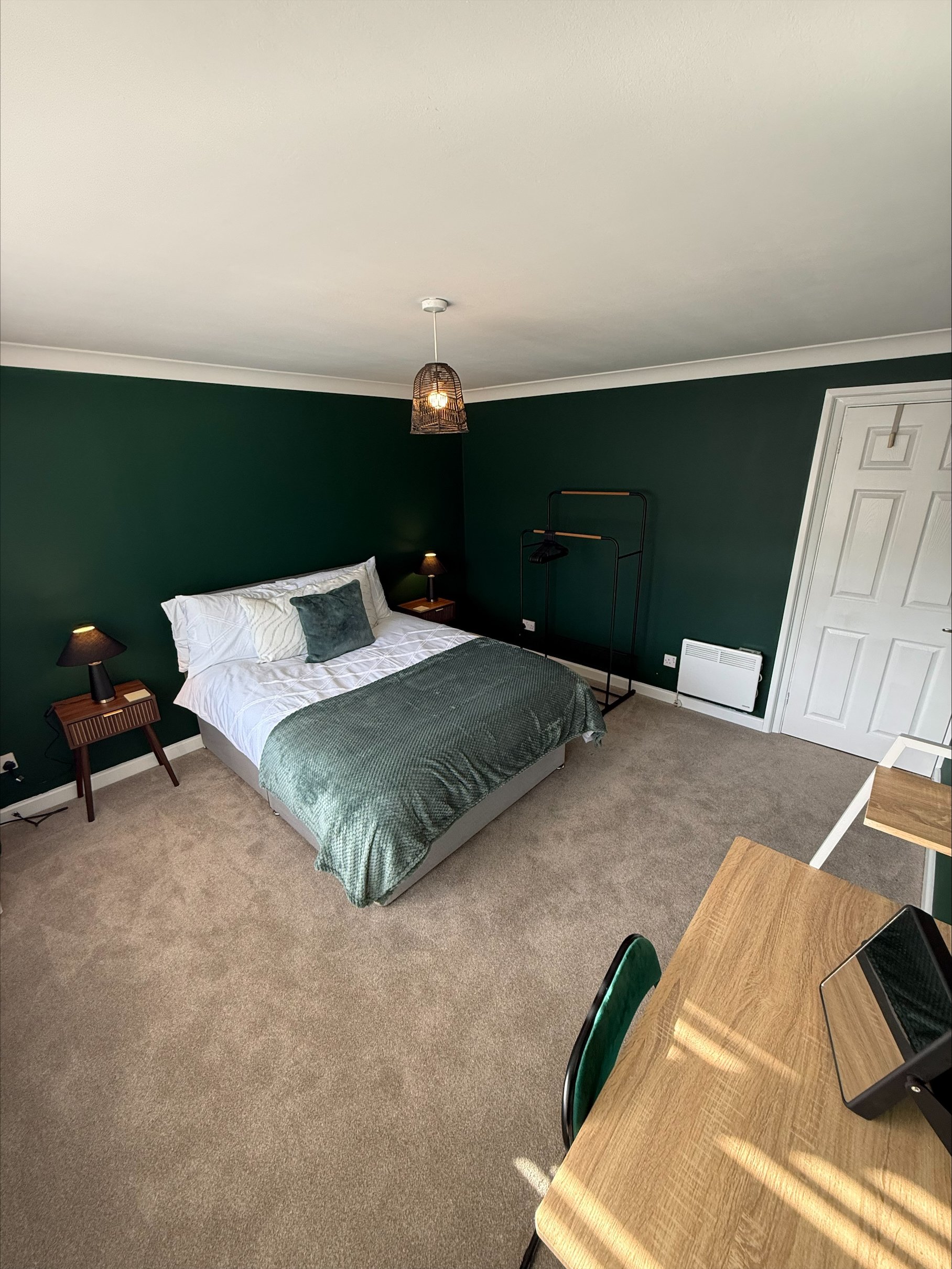 Modern bedroom with dark green walls, neatly made bed, woven pendant light, bedside lamps, clothing rack, and wooden desk.